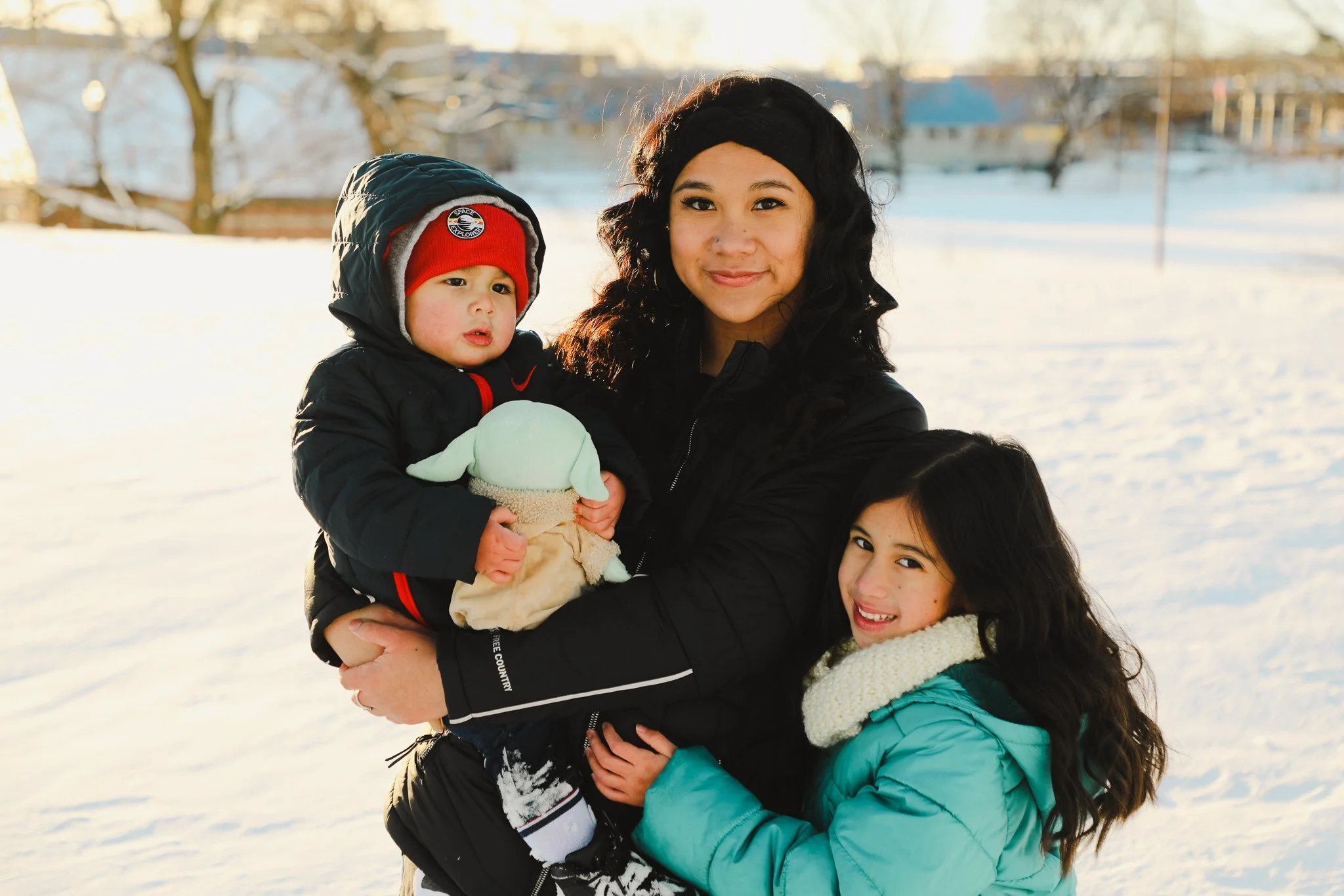A woman with black curly hair smiling and holding a young child dressed in a black coat and red hat, and a teenage girl in a turquoise coat with a white scarf, standing outdoors in a snowy landscape during daytime.