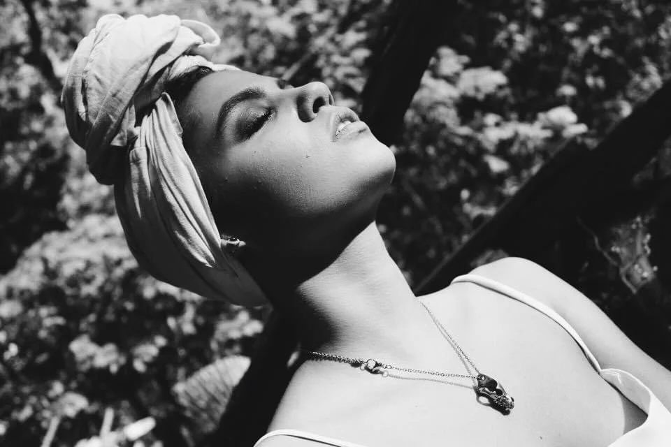 A woman with a headwrap and necklace, her eyes closed, head tilted back against a natural outdoor background, in black and white.