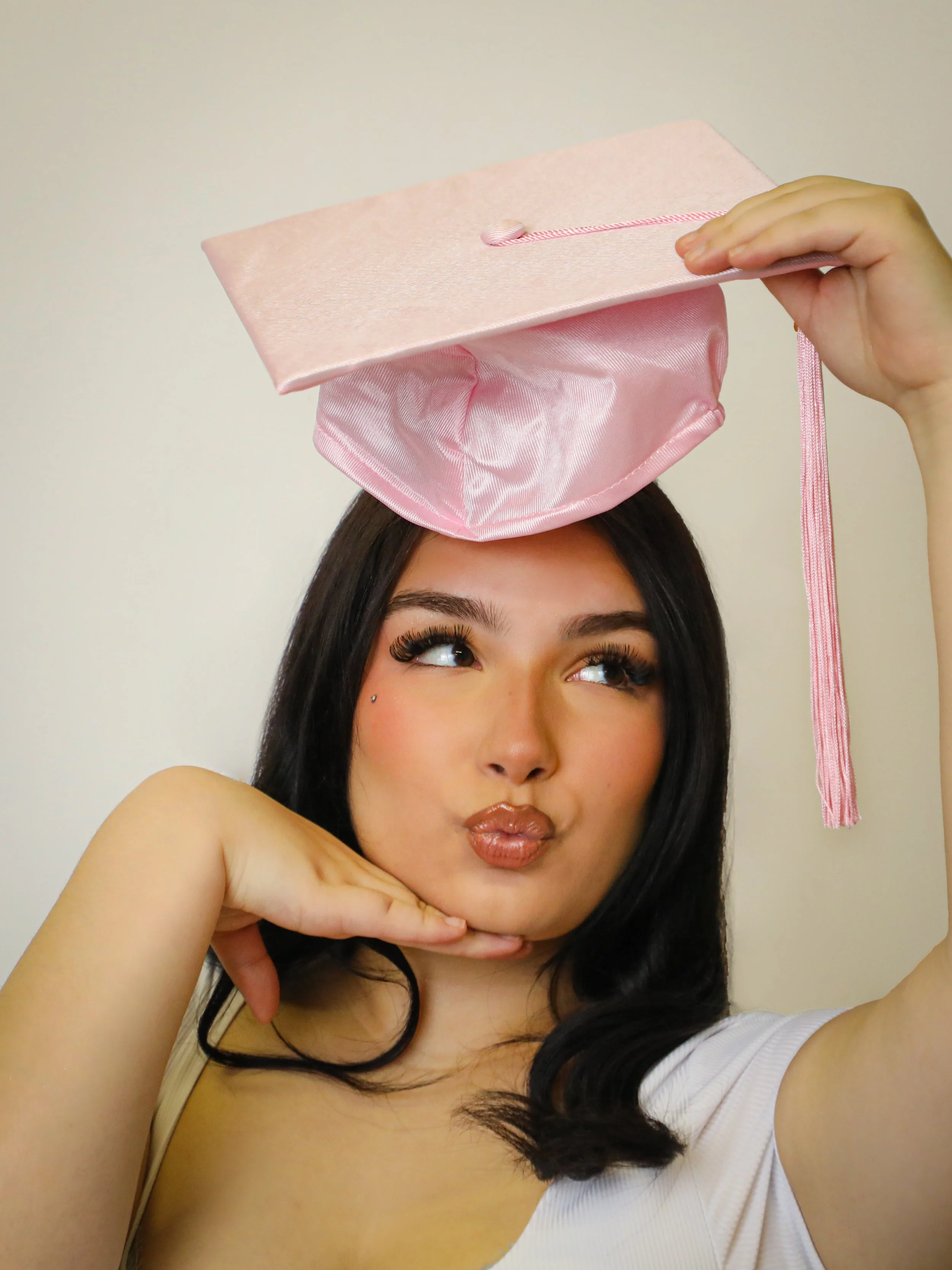 A woman with long dark hair, makeup, and a white top, balances a pink graduation cap on her head, with her lips pursed and her hand under her chin, against a plain background.