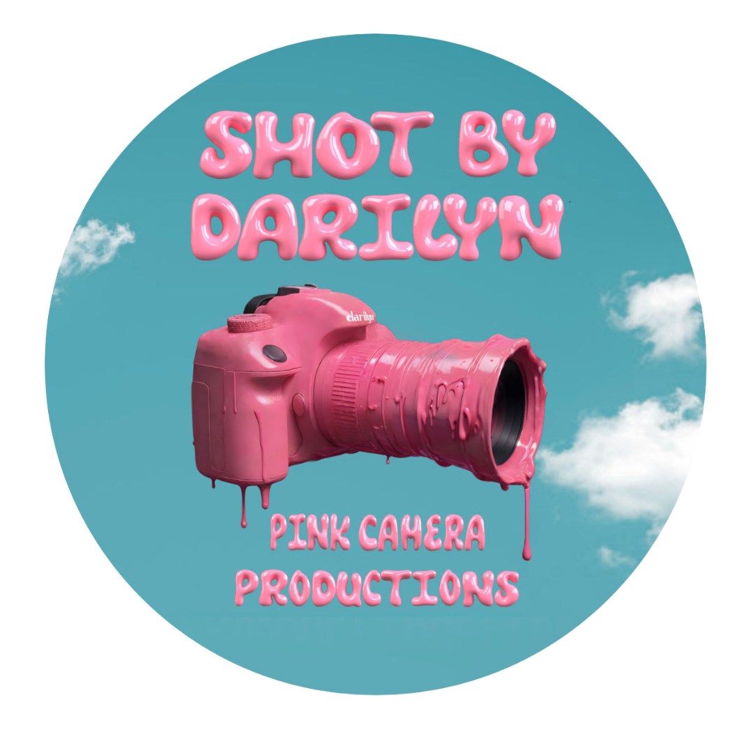 A pink camera with a dripping paint effect is centered against a blue sky background with clouds. The text above the camera reads "Shot by Darilyn" in pink, puffy lettering. Below, the text reads "Pink Camera Productions," also in pink, puffy lettering.
