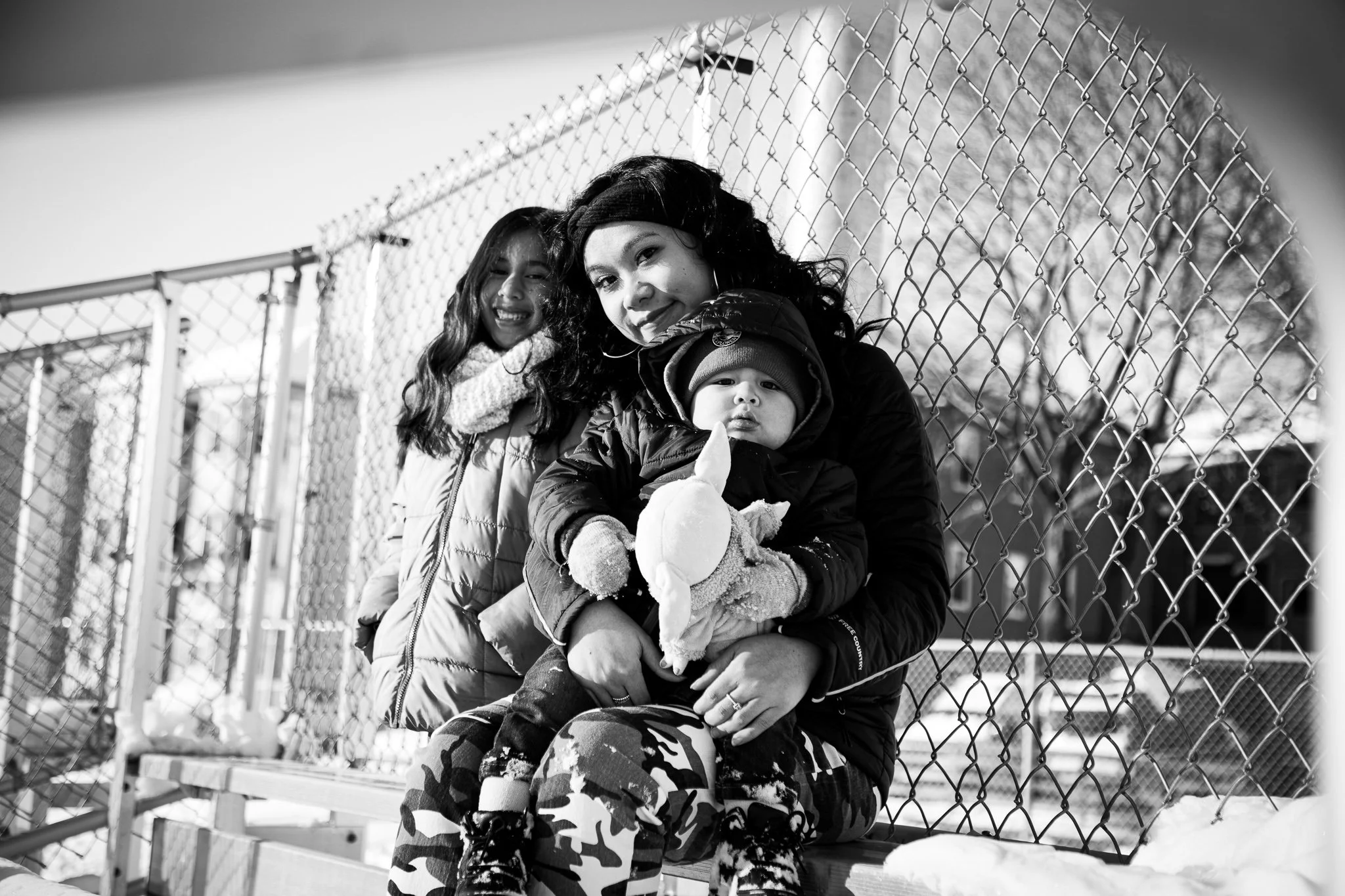 A woman with long curly hair holding a young child with a stuffed animal, sitting on a bench outside near a chain-link fence. Behind them, a girl with long hair and a scarf is smiling. Snow is visible on the ground.