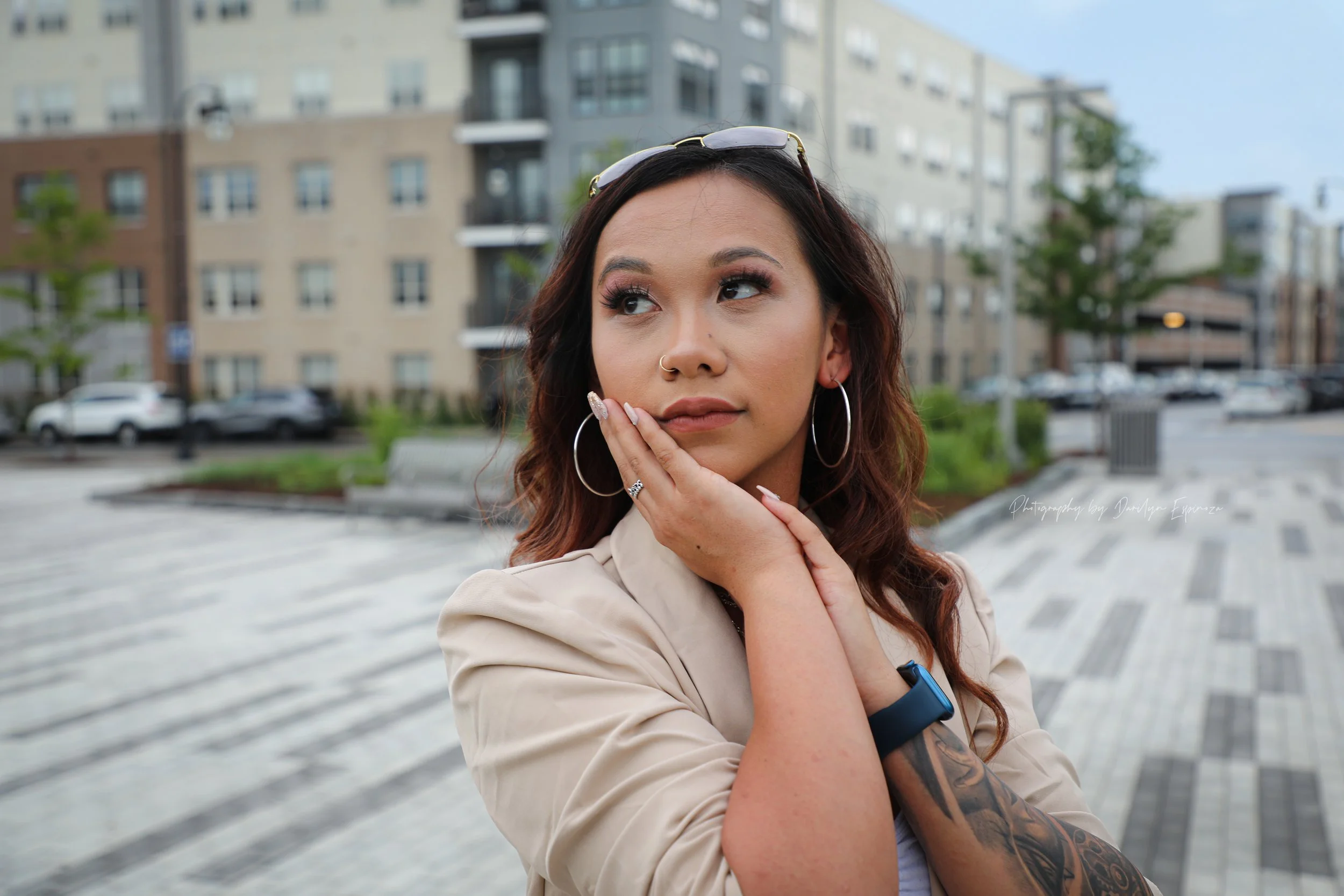 A young woman with long wavy hair, wearing hoop earrings and a beige blazer, stands outdoors in an urban setting with modern buildings, parked cars, and trees in the background. She has a thoughtful expression, with her hand resting on her chin, and 