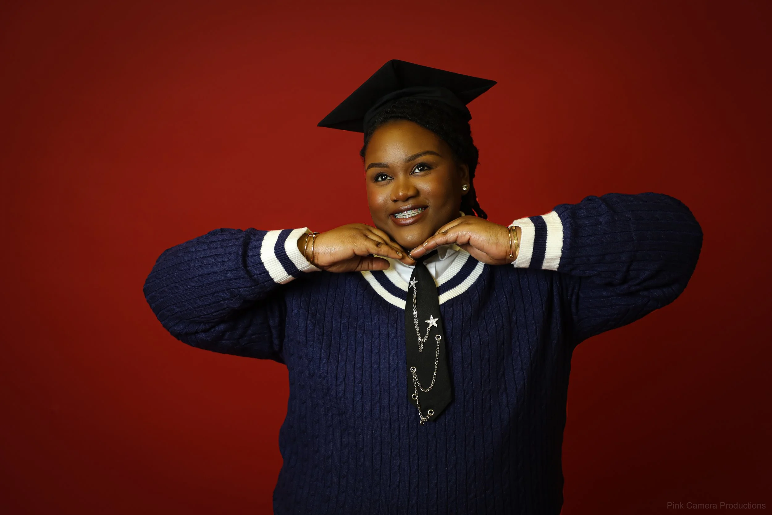 A woman in a blue sweater and graduation cap poses with her hands near her chin against a red background.