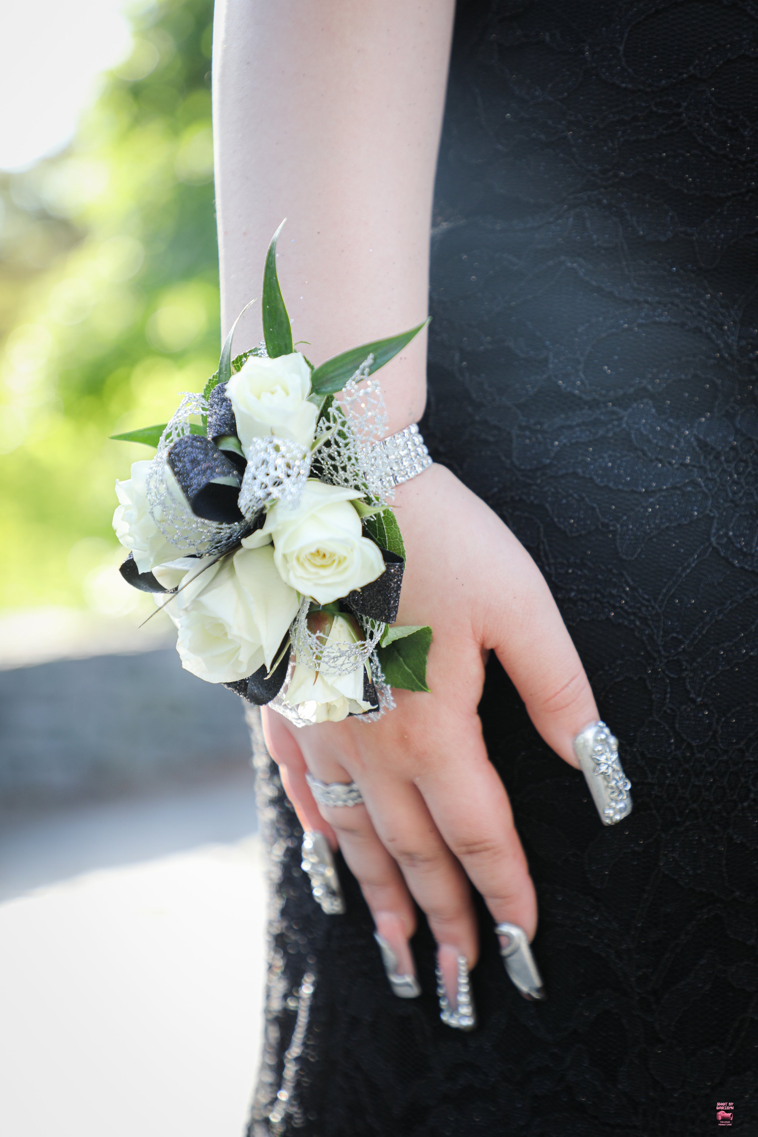 Close-up of a hand with silver and diamond rings, wearing a silver bracelet, and a corsage of white roses, black ribbons, and green leaves on the wrist, against a blurred outdoor background.