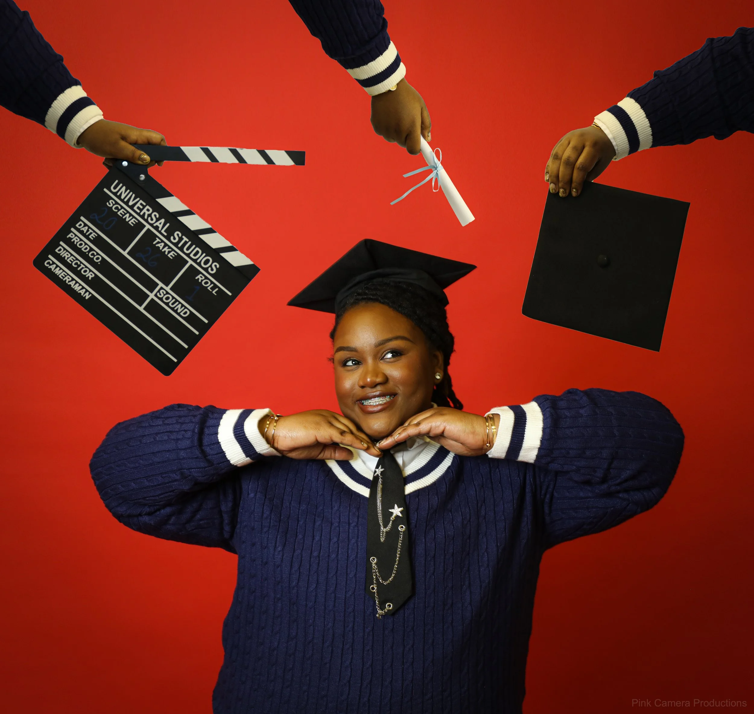 A young woman in a graduation cap and navy blue sweater poses with her hands under her chin, smiling. Around her, there are hands holding a film clapperboard, a rolled diploma, and a mortarboard against a red background.