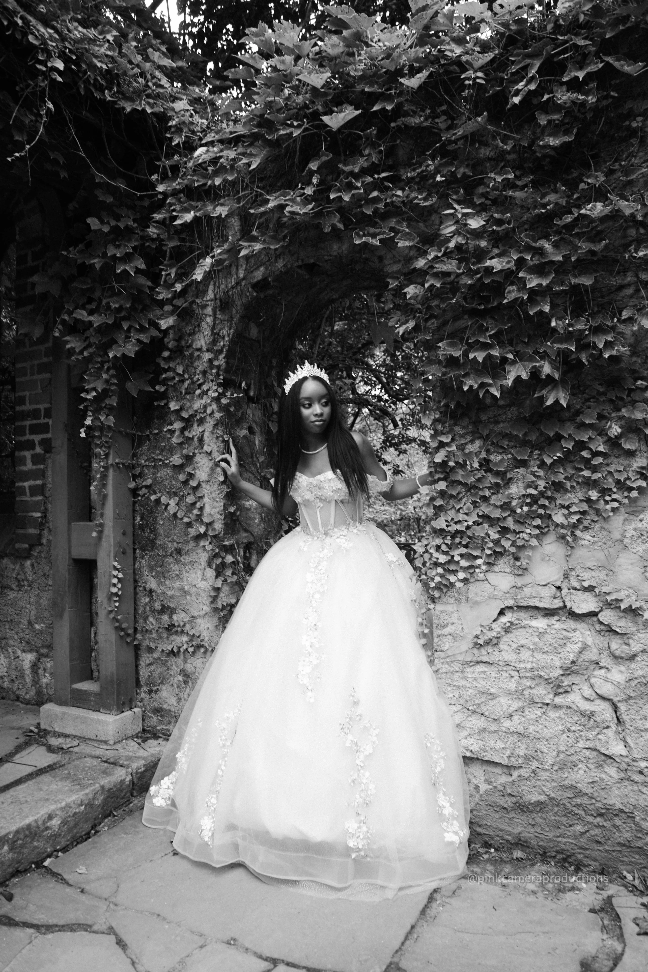 A woman in a wedding dress standing in front of an archway covered in vines and leaves, in black and white.