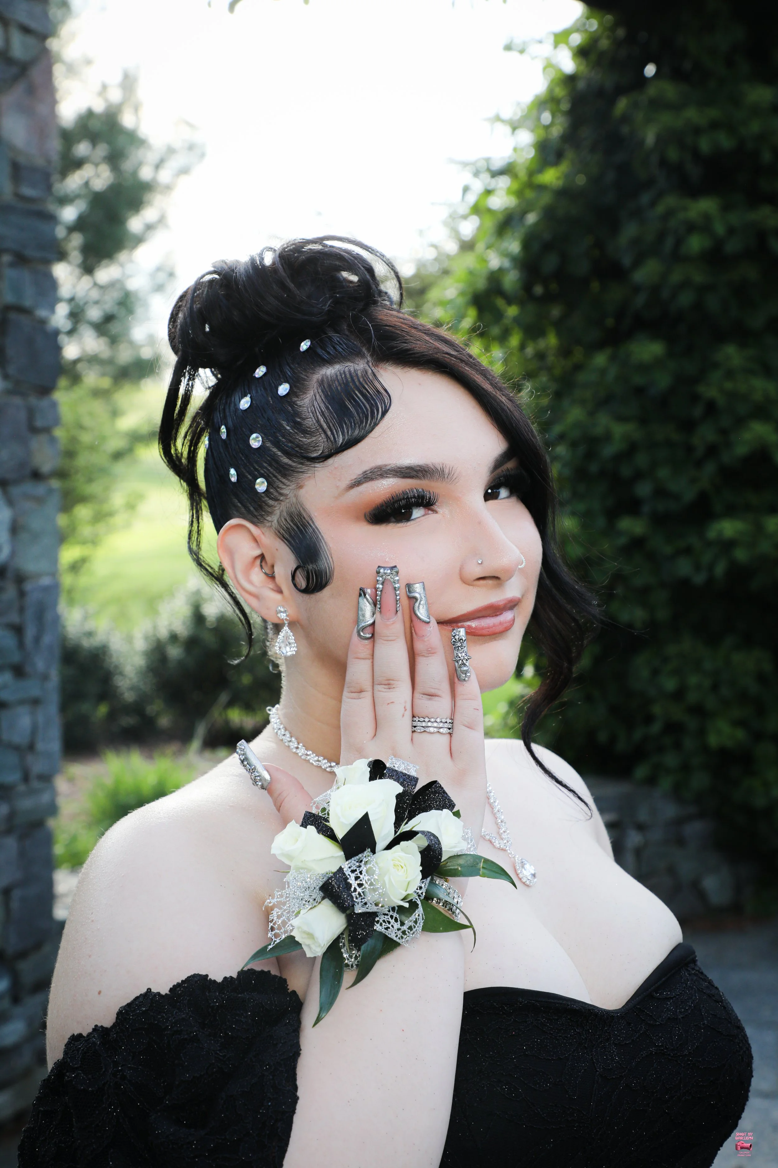 A woman with dark hair styled in an elaborate updo, adorned with rhinestones, wearing diamond jewelry, a floral corsage on her wrist, and a black dress, smiling outdoors with greenery in the background.