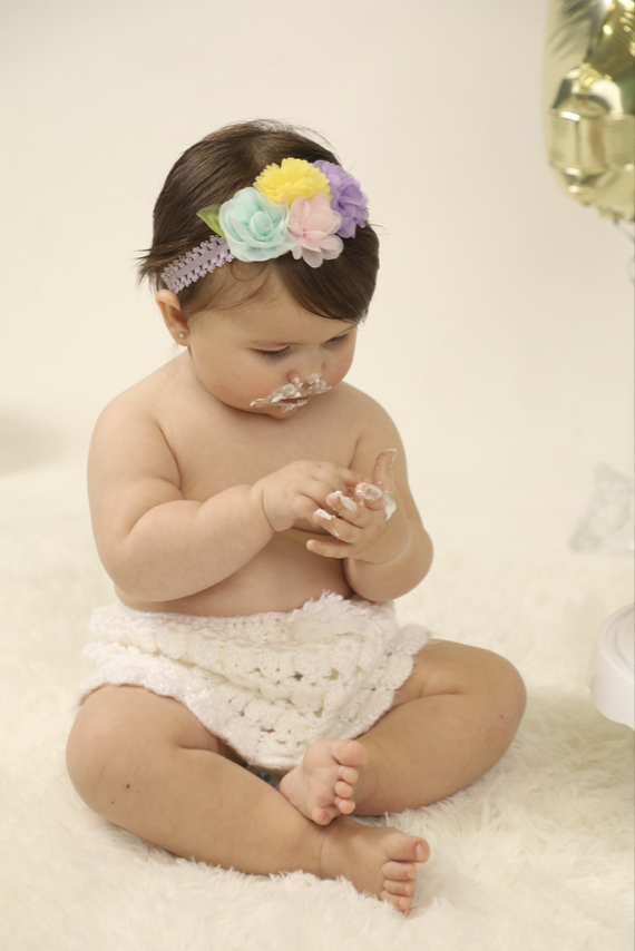 A baby girl with a floral headband, sitting on a soft white rug, playing with white foam or cream, with a cream-colored background.
