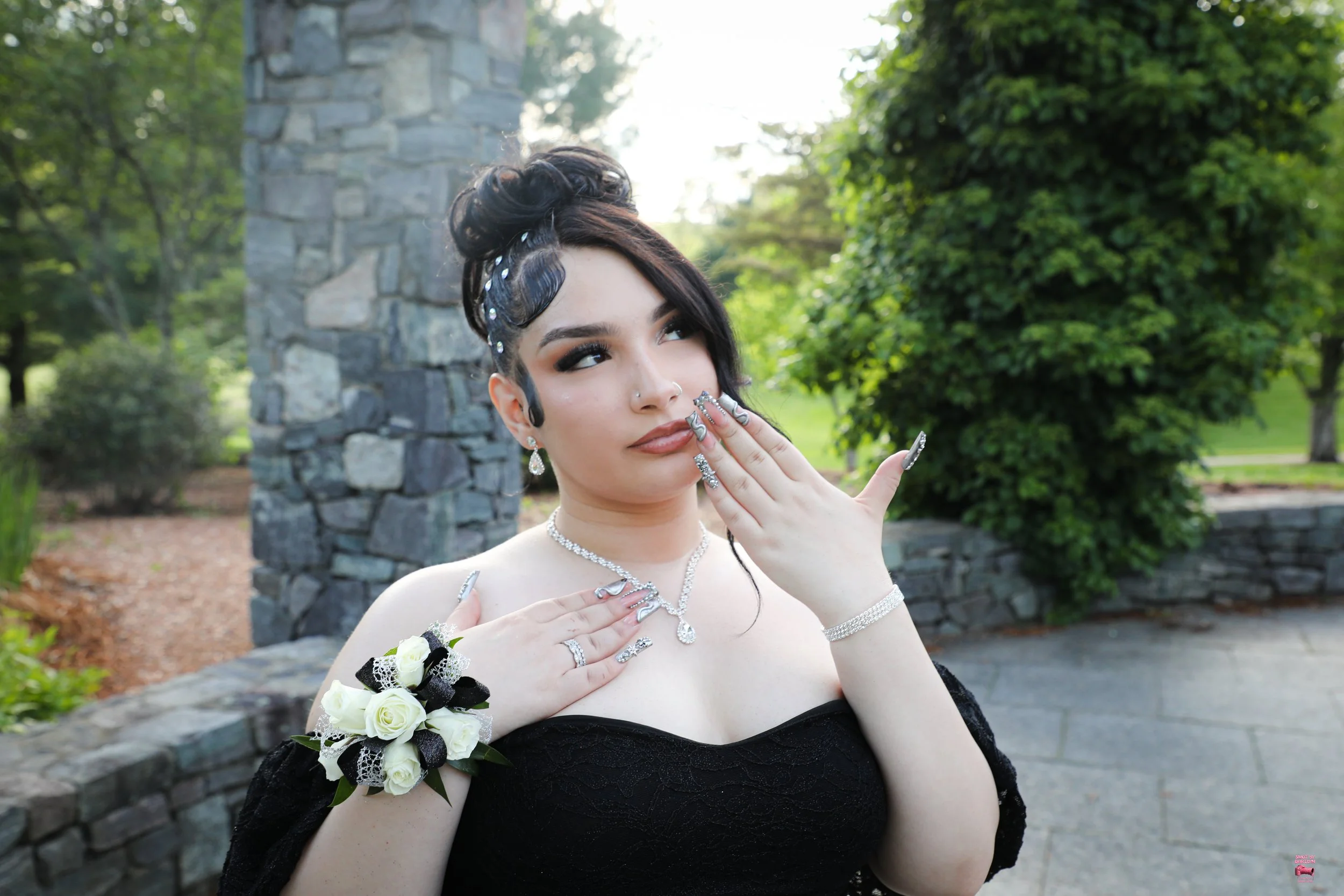 A woman in a black dress with jewelry and a floral corsage, standing outdoors near a stone structure and greenery, with her hands near her face in a thoughtful pose.