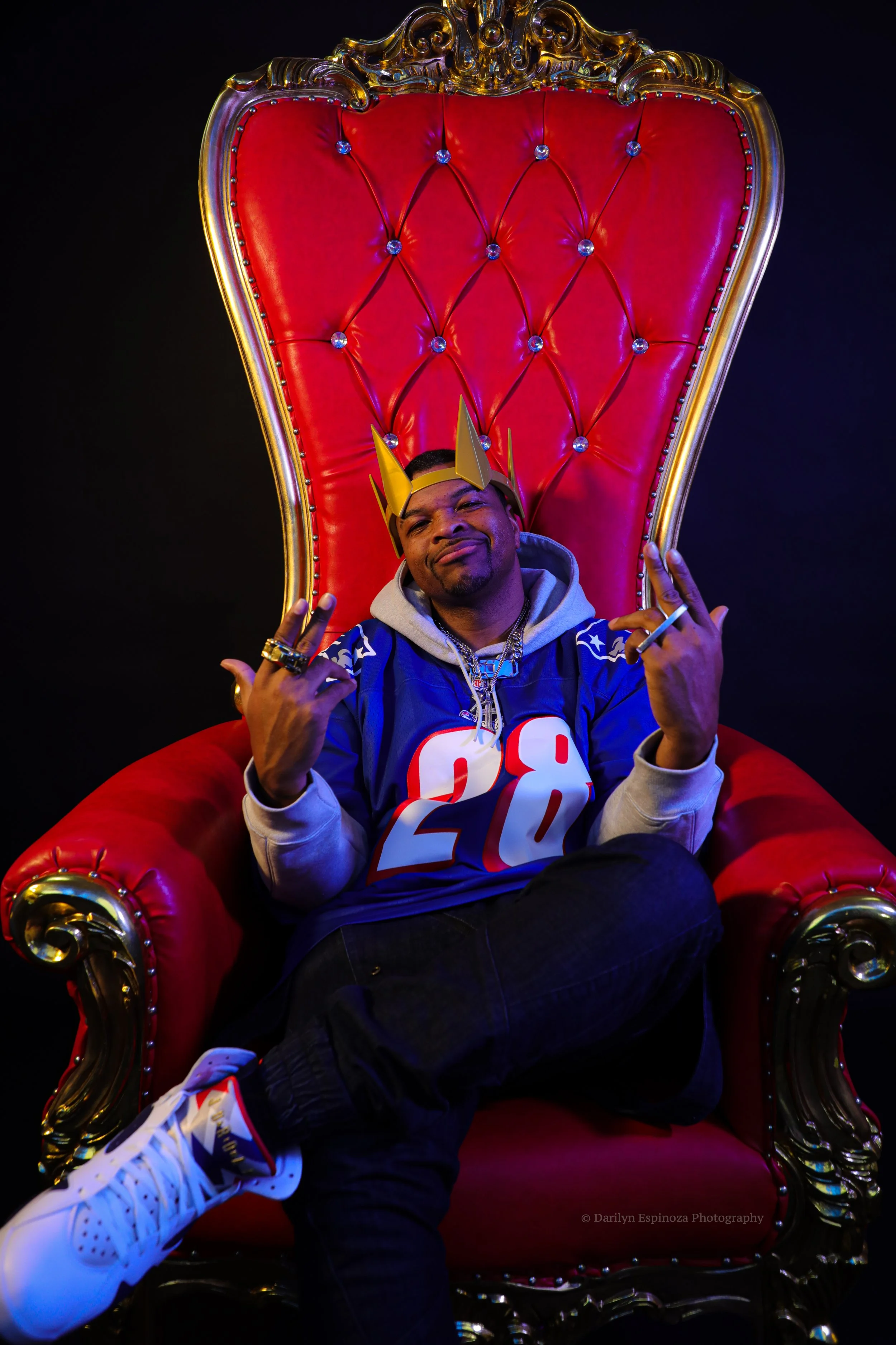 A man sitting on a red, ornate throne-like chair with gold accents, wearing a blue sports jersey, gray hoodie, white sneakers, a crown, and accessories, striking a confident pose.