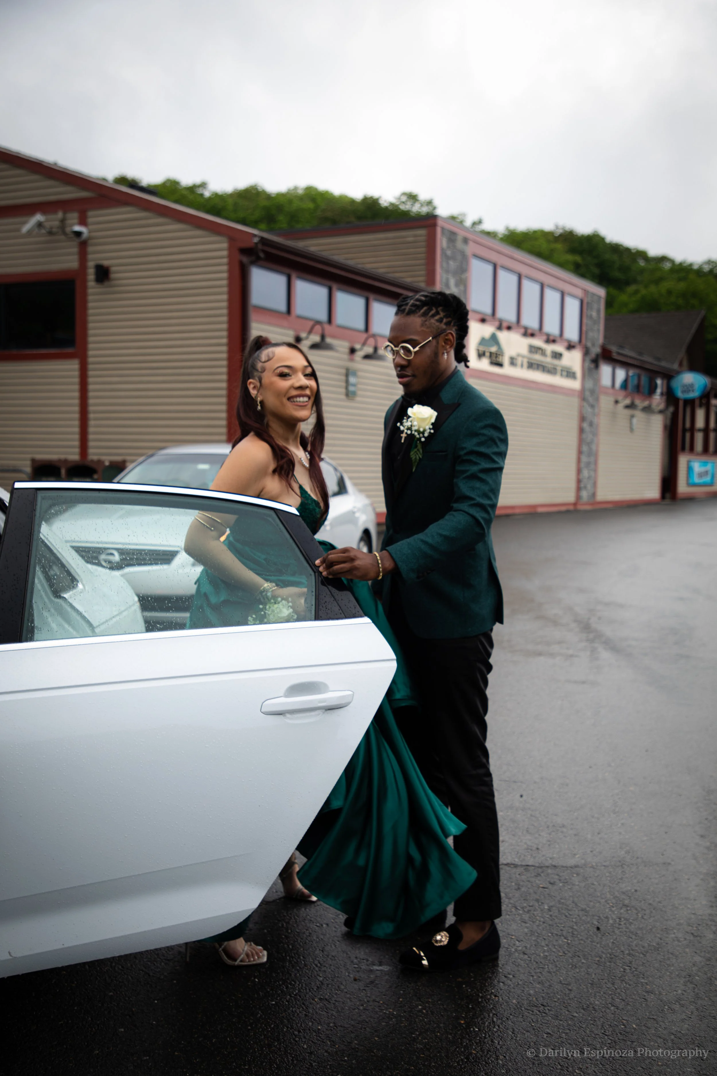 A couple dressed in formal attire standing outside a white car on a wet pavement, with a building in the background, likely celebrating a special occasion.