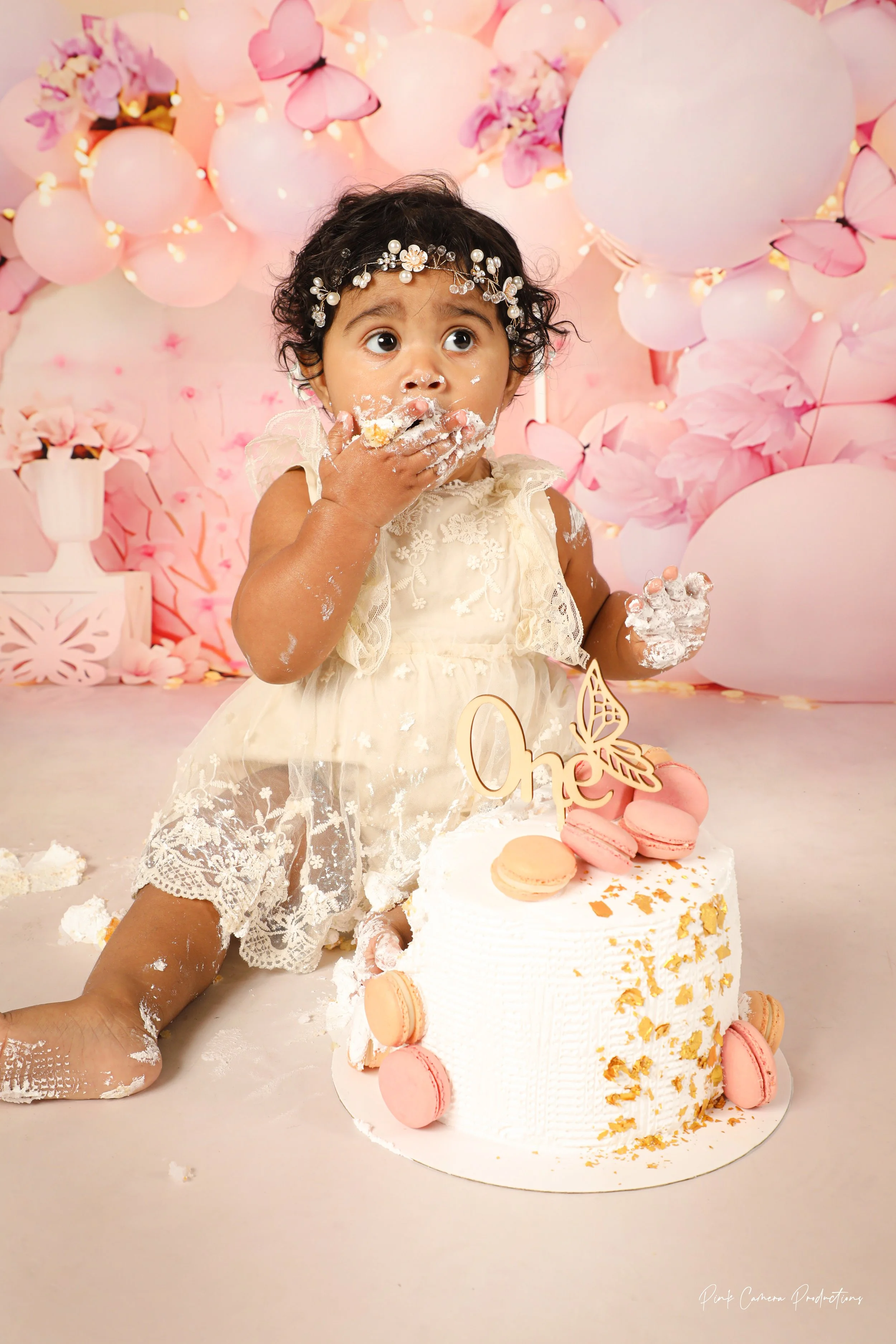 A young girl in a cream lace dress with a pearl and floral headband eats cake at a first birthday celebration. The cake is decorated with pink macarons and gold accents, and a 'One' cake topper. The background features pink balloons and flowers.