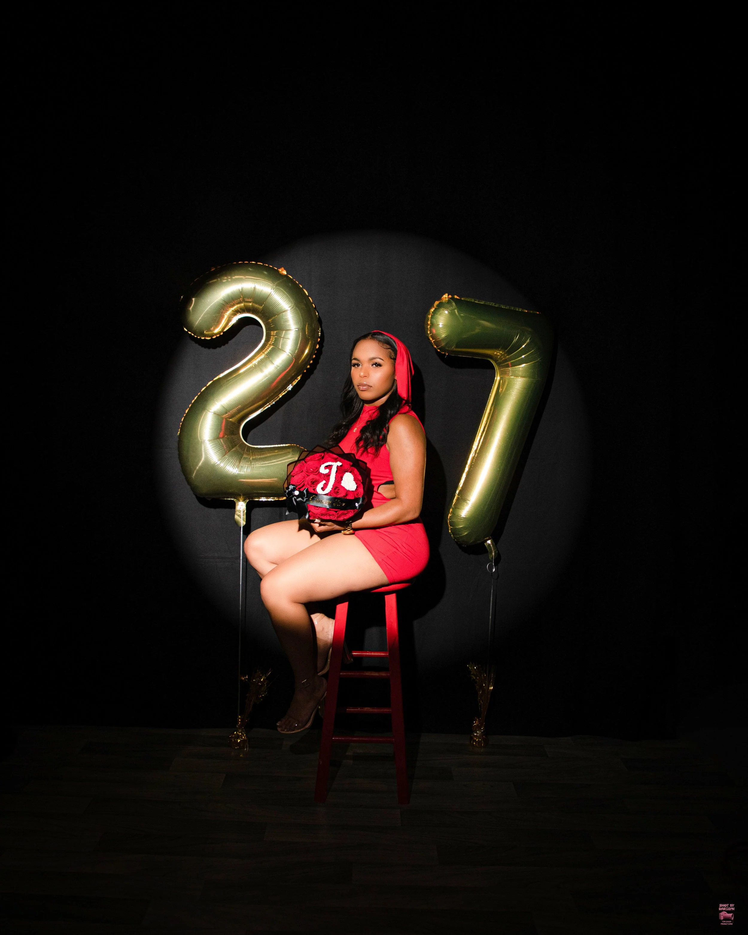 A woman dressed in a red outfit sitting on a red stool with large gold balloon numbers '2' and '7' behind her, holding a bag with a red bow and initials; dark background with spotlight on her.