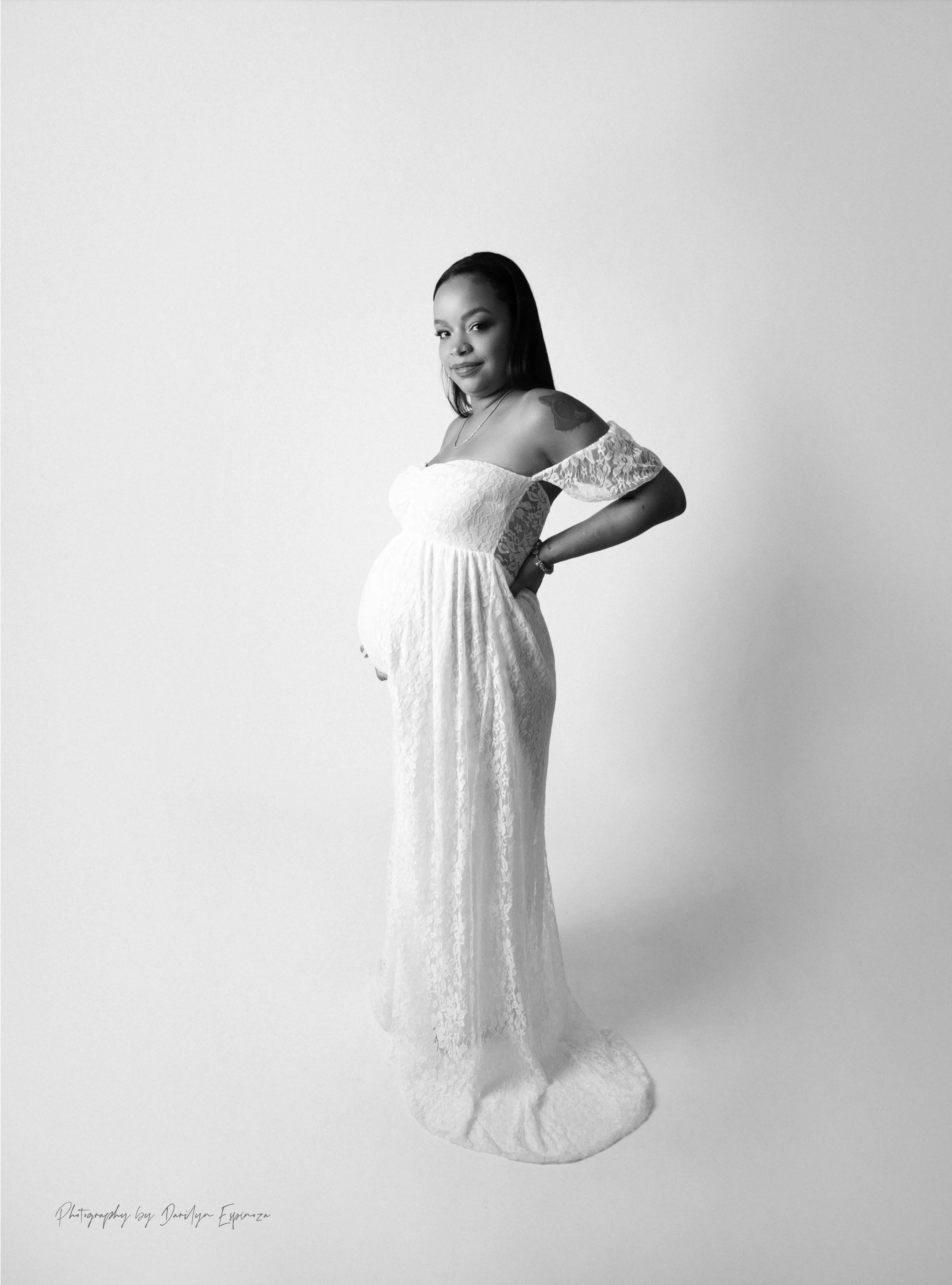 Black and white portrait of a pregnant woman in a long lace dress, standing against a plain light background, with her hand on her hip, looking at the camera.