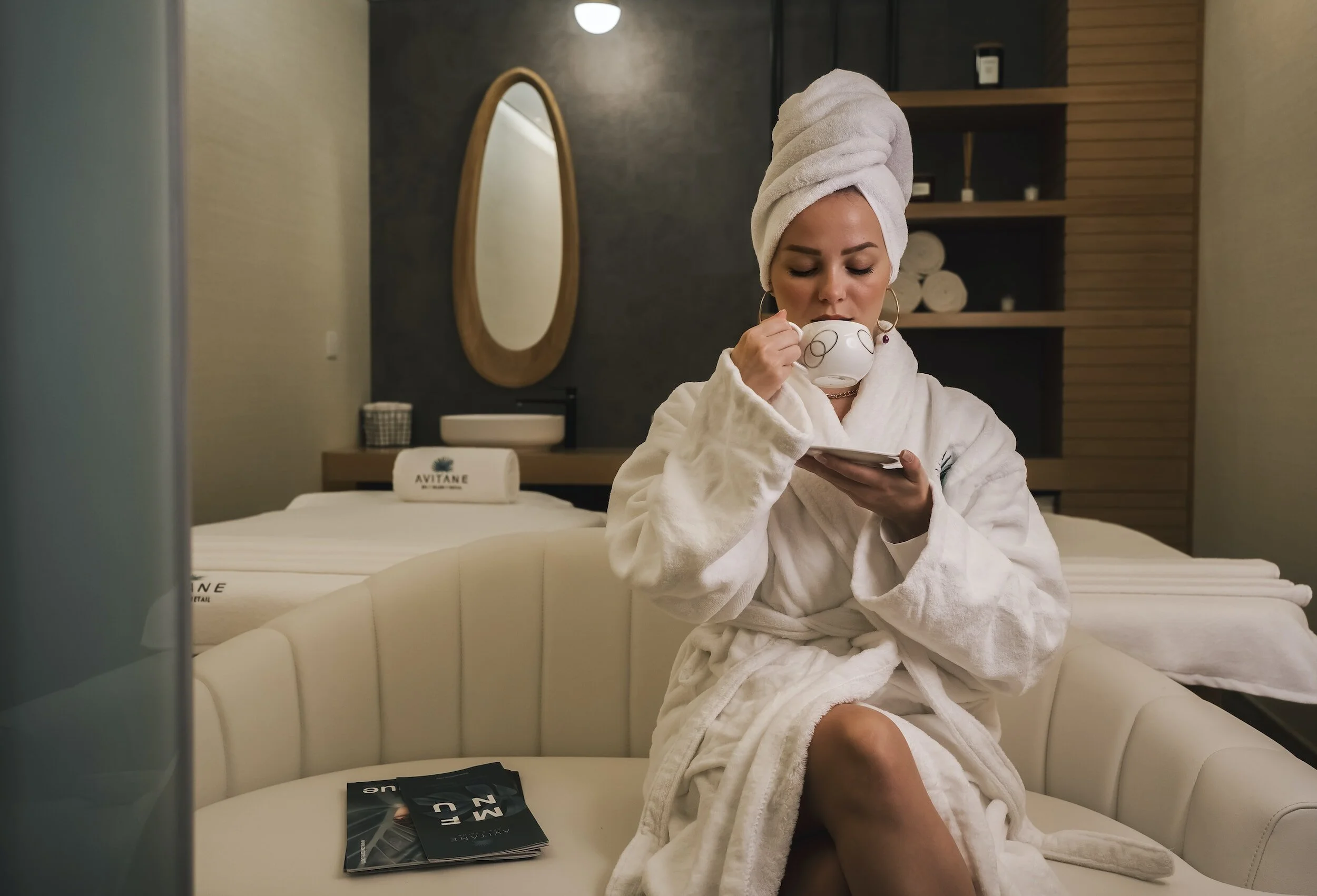 A woman in a bathrobe and towel wrapped on her head, sitting on a cream-colored couch, drinking from a cup with a book on her lap in a cozy hotel room.