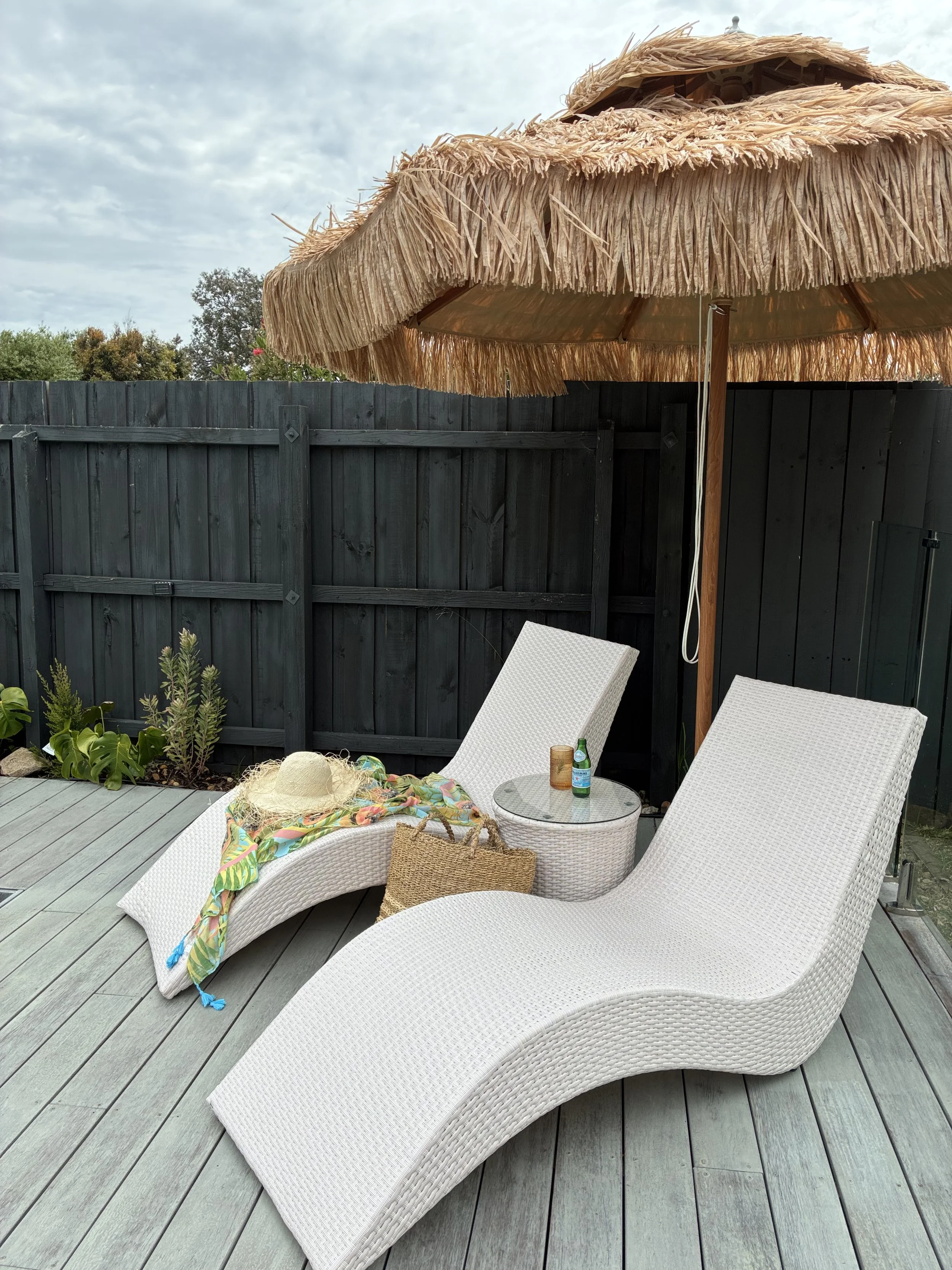 Two white wicker lounge chairs on a wooden deck, one with a colorful beach towel, a sun hat, and a woven bag, beside a small round glass table with drinks, under a large straw umbrella, with a black fence and some plants in the background.