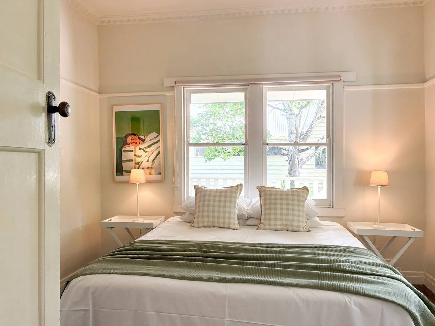 Country-style holiday home bedroom with a sage green throw, gingham cushions, bedside lamps and artwork