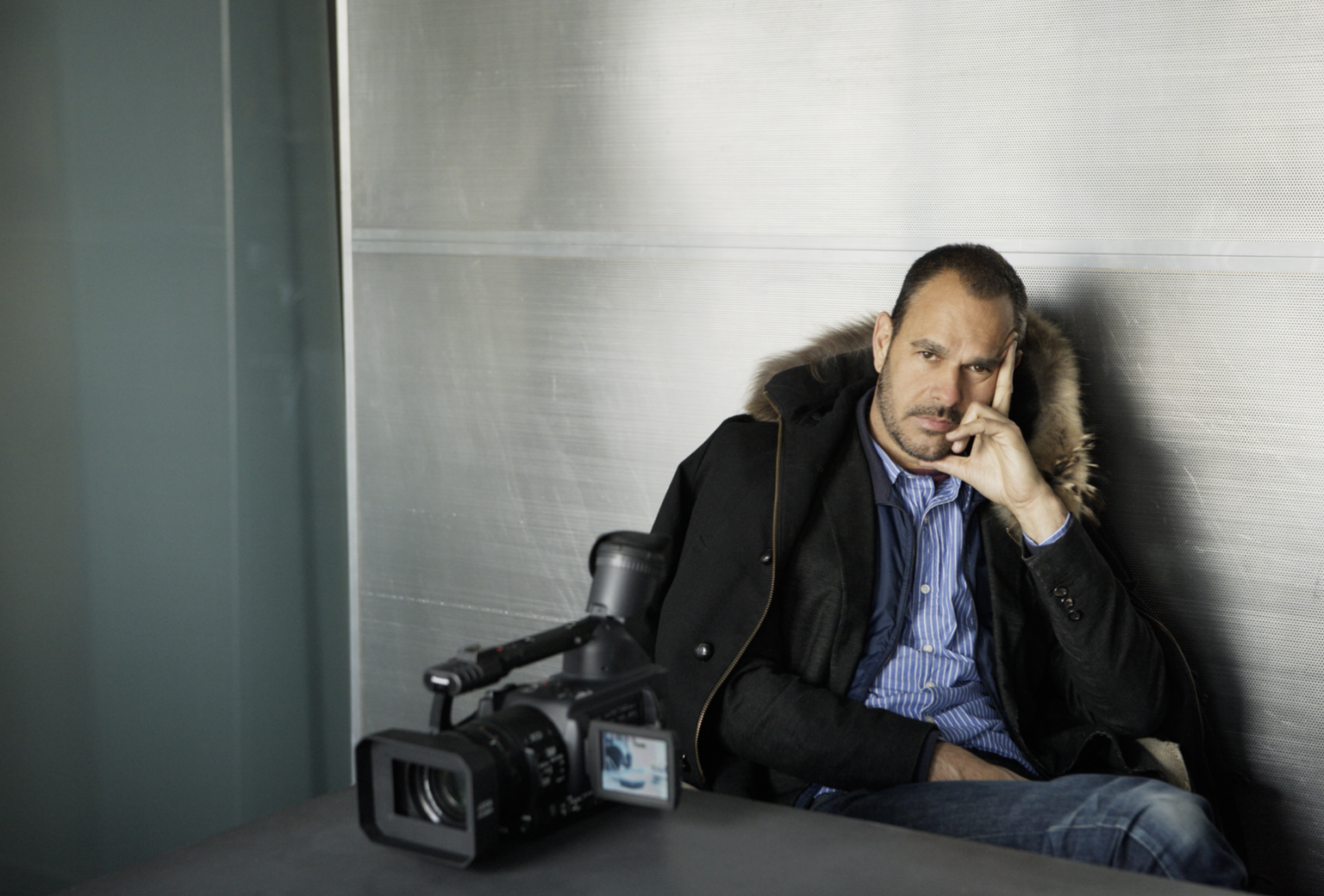 A man sitting and posing, wearing a fur lined coat over a suit jacket, blue stripped button up and jeans. A video camera is on the table.