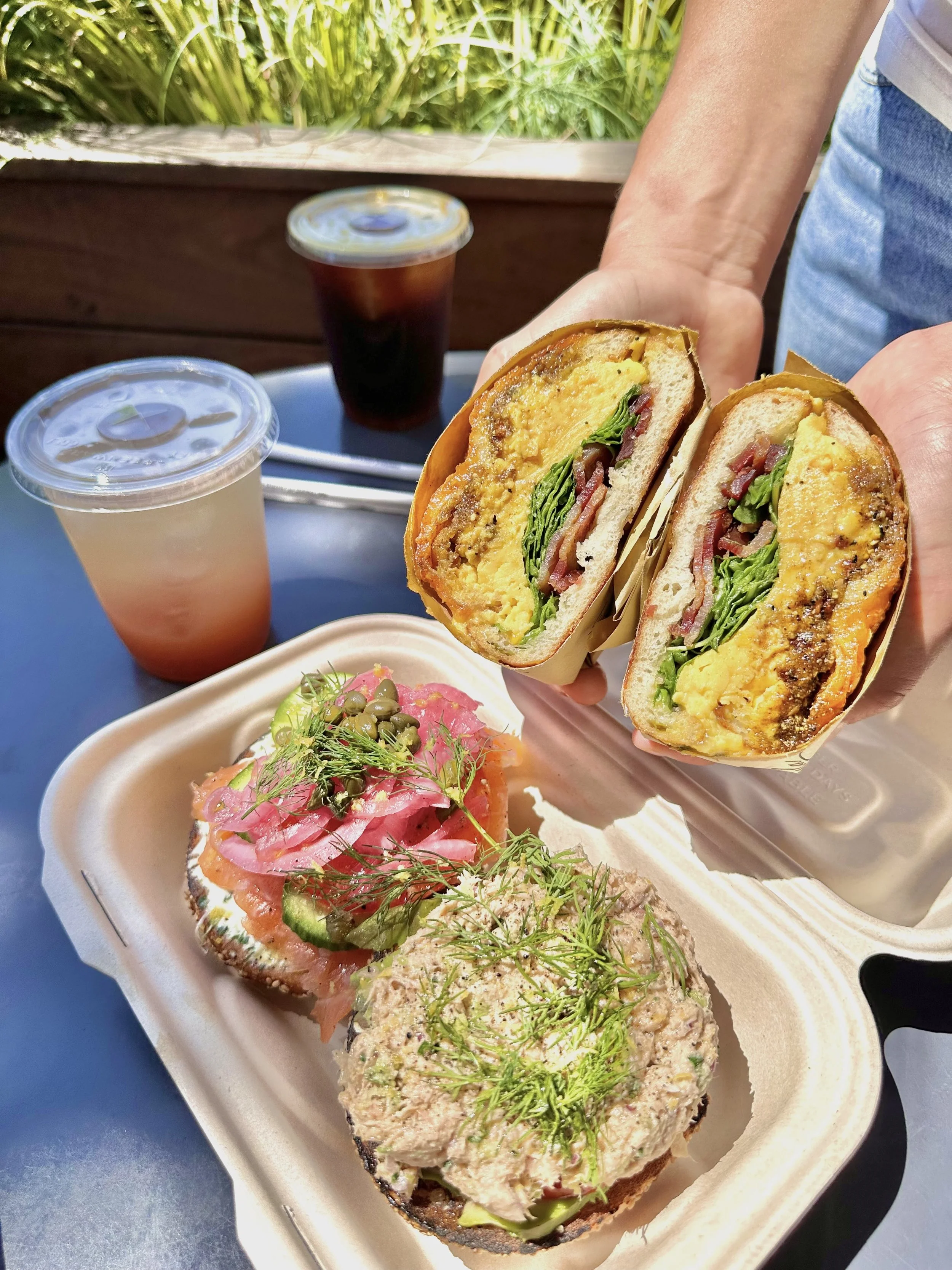 Woman holding a Layla bagel split in half above a box of the Laika bagel and Tuna Melt bagel with an arnold palmer lemonade in the background