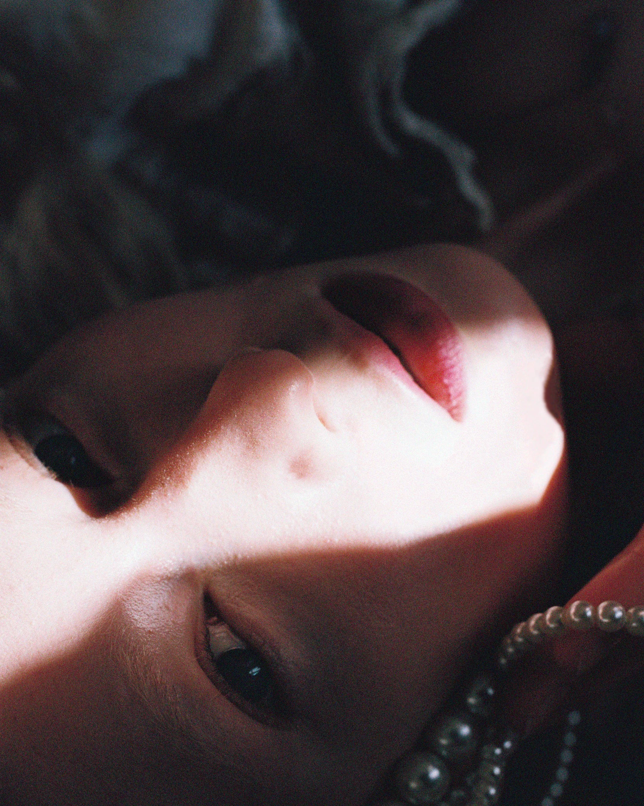 Close-up of a woman with pink lipstick and dark nail polish, wearing pearl necklaces.