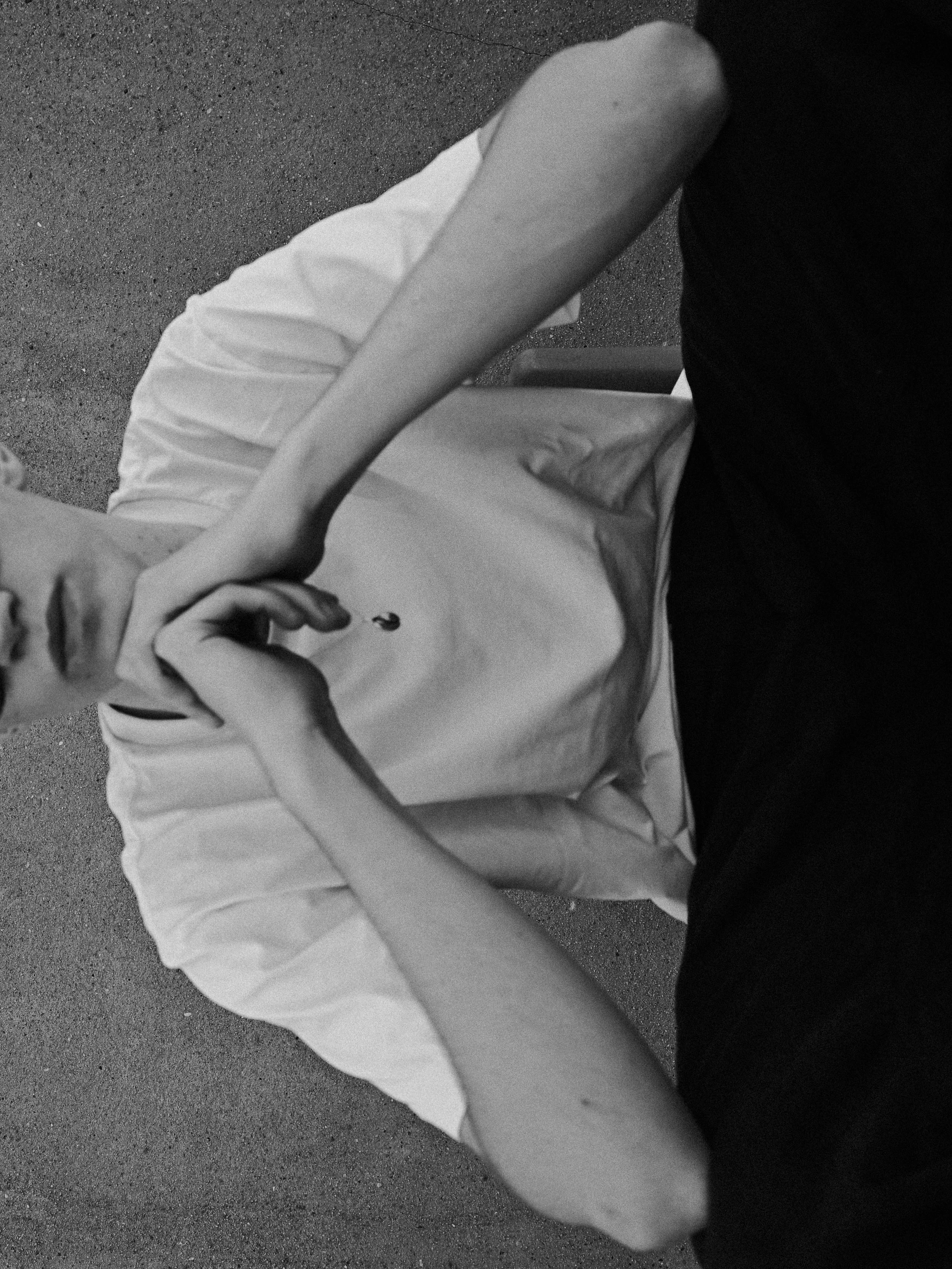 Close-up of a person sitting on a chair with a white tablecloth, wearing a dark shirt, on a textured outdoor surface, in black and white.