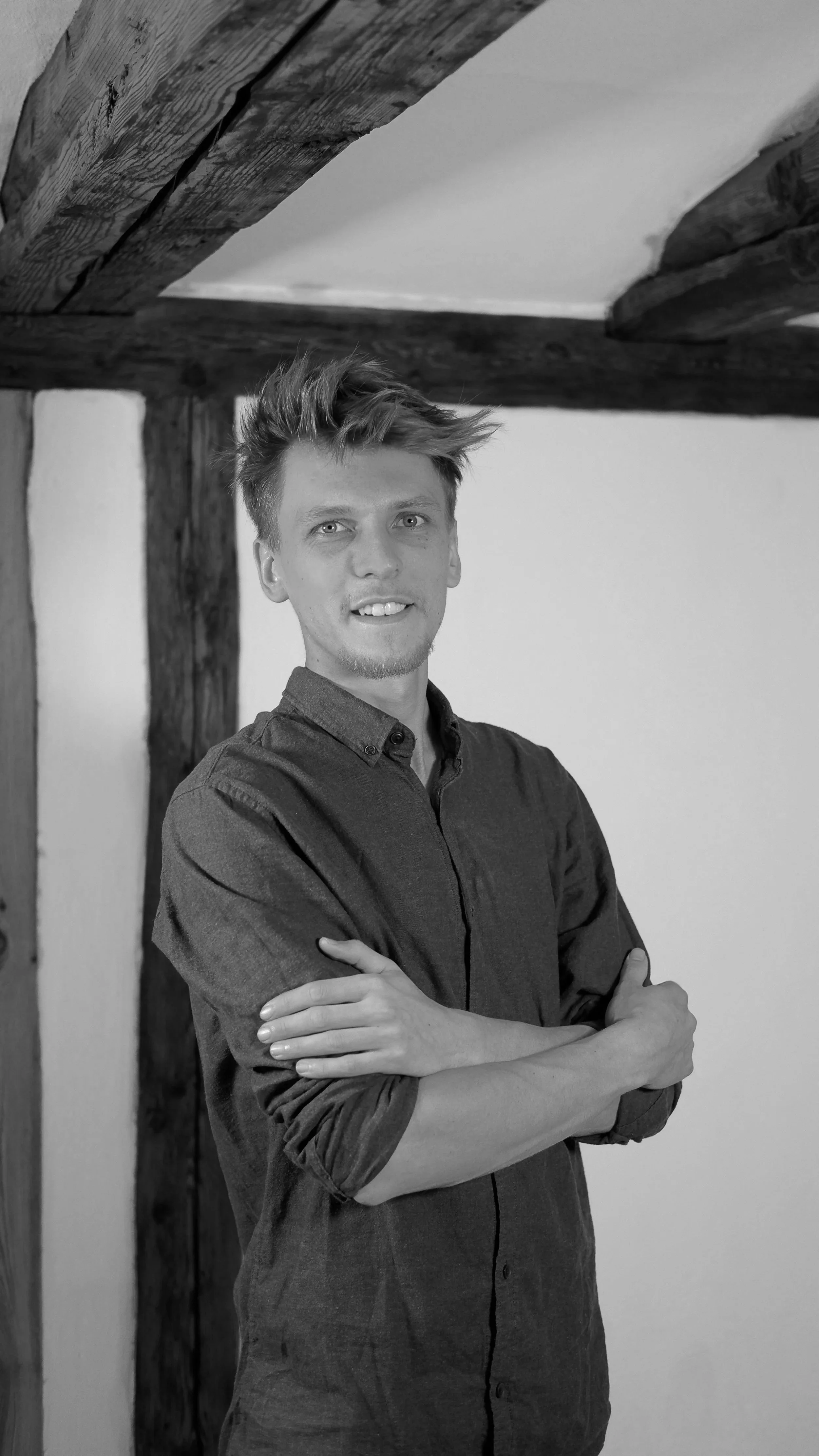 Schwarz-Weiß-Fotografie eines jungen Mannes, der lächelnd mit verschränkten Armen vor einer Wand steht, im Hintergrund sichtbar Holzbalken.