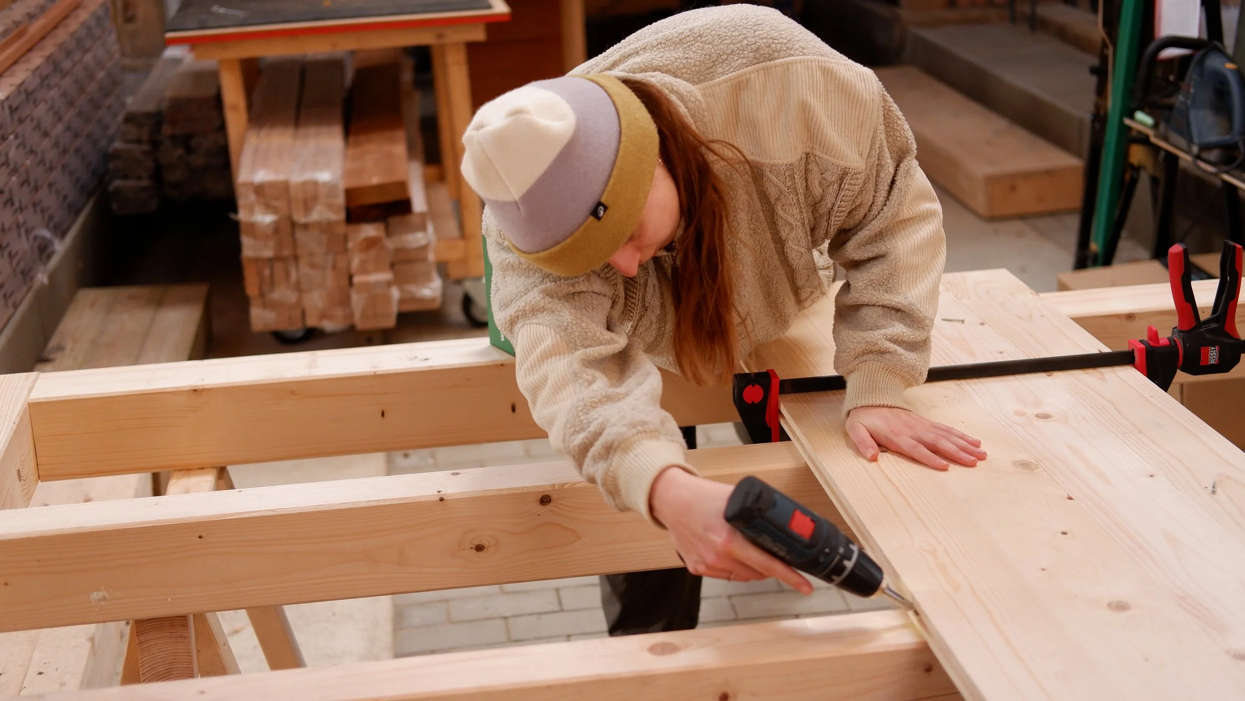 Ein Mensch arbeitet in einer Werkstatt an einem Holzbearbeitungsprojekt, benutzt eine Bohrmaschine, trägt warme Kleidung und eine Mütze.
