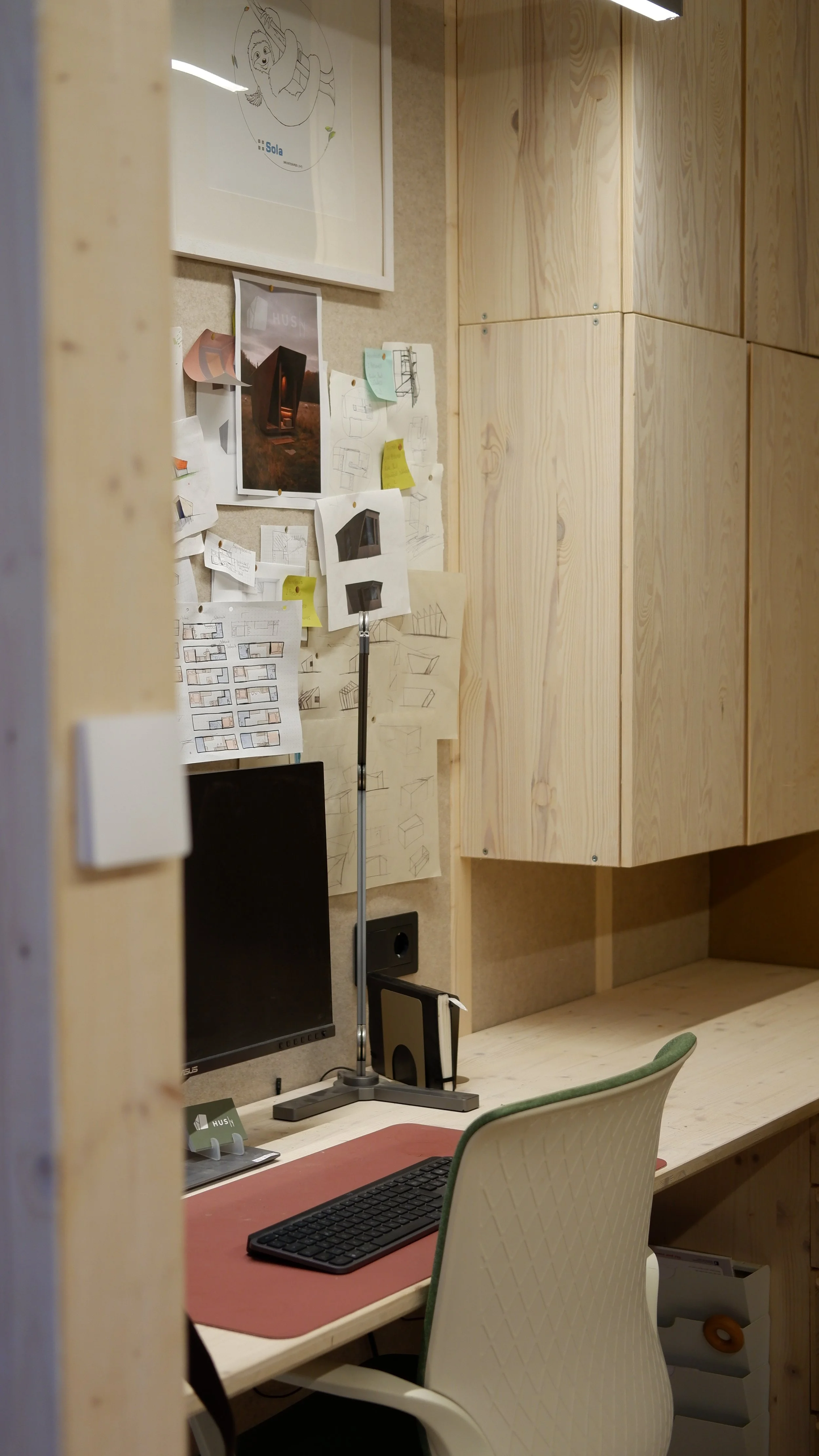 Ein Büroarbeitsplatz mit einem weißen Stuhl, einem Computerbildschirm, Tastatur, Mauspad und einer Wand mit angebrachten Skizzen, Farbdrucken und Notizen. Es gibt auch eine Holzregalwand im Hintergrund.