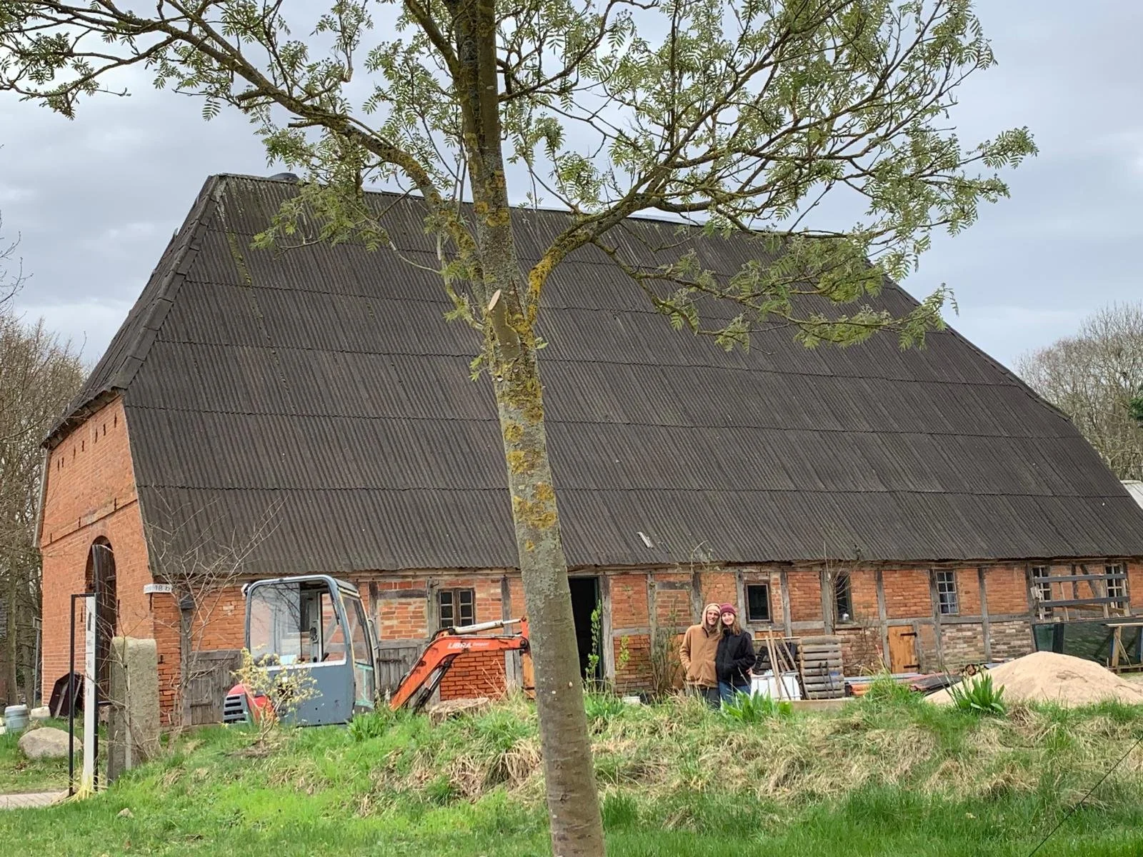 Ein großes Schulmaß, das auf einem Feld steht, neben einem Baum. Dahinter ein rot-verschiefertes Gebäude, das gerade renoviert oder gebaut wird. Zwei Menschen stehen vor dem Gebäude und lachen. Es gibt Handwerkergeräte und Baumaterialien im Vordergrund.