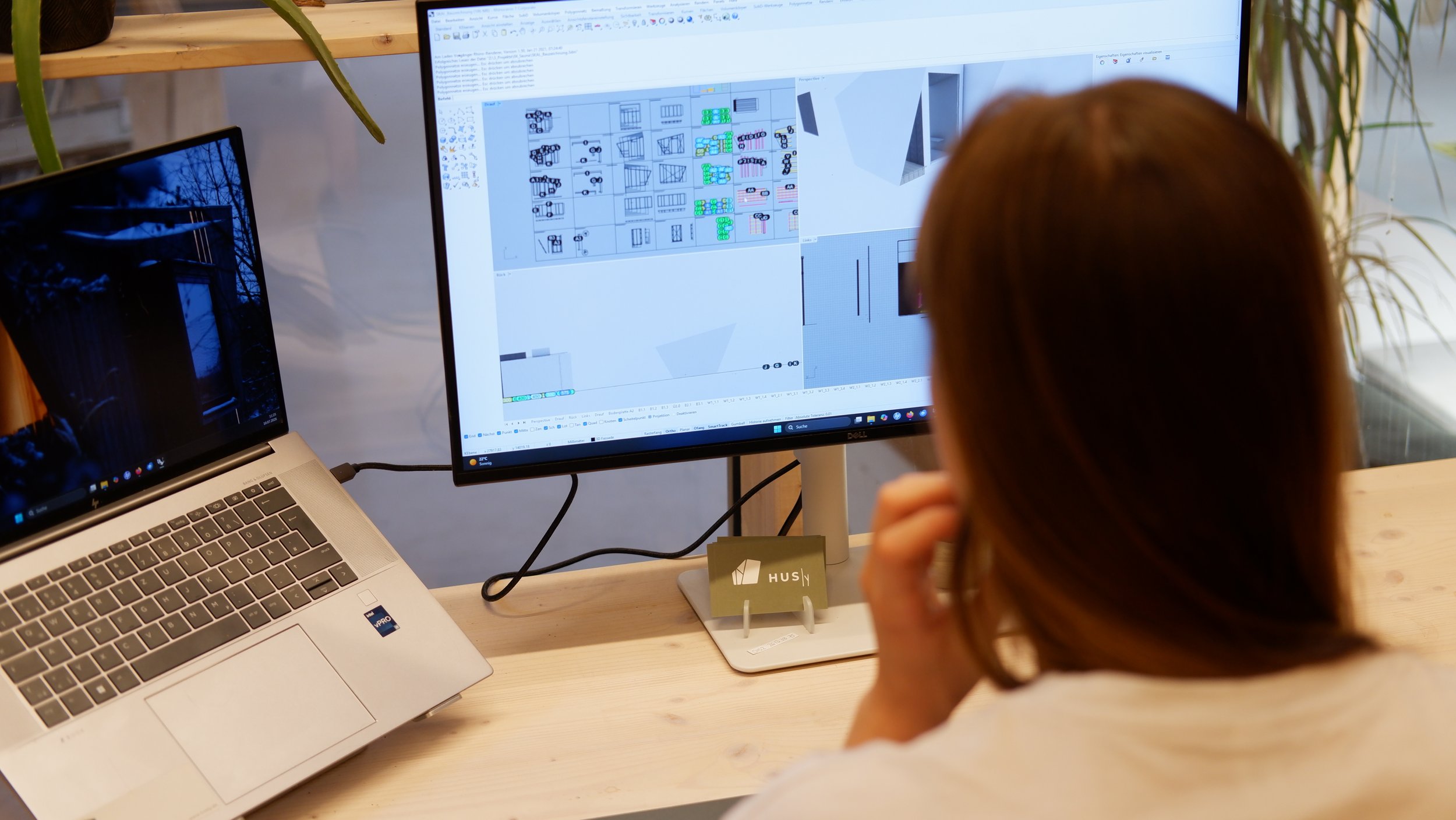 Eine Person arbeitet an einem Computer mit einem großen Bildschirm, auf dem Architekturzeichnungen und Pläne zu sehen sind, in einem Raum mit Pflanzen und einem Laptop auf einem Holztisch.