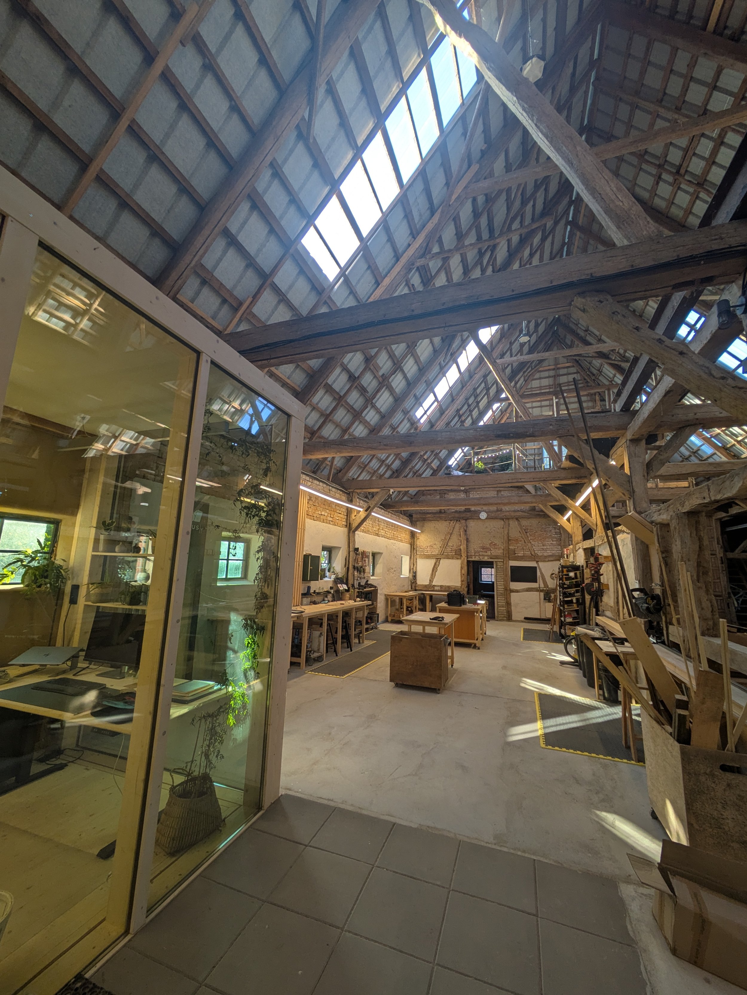 Blick in einen modernen, industriell gestalteten Co-Working Space mit Holzdecke, großen Fenstern und Arbeitsplätzen. Es gibt Tische, Stühle, Pflanzen und Regale.