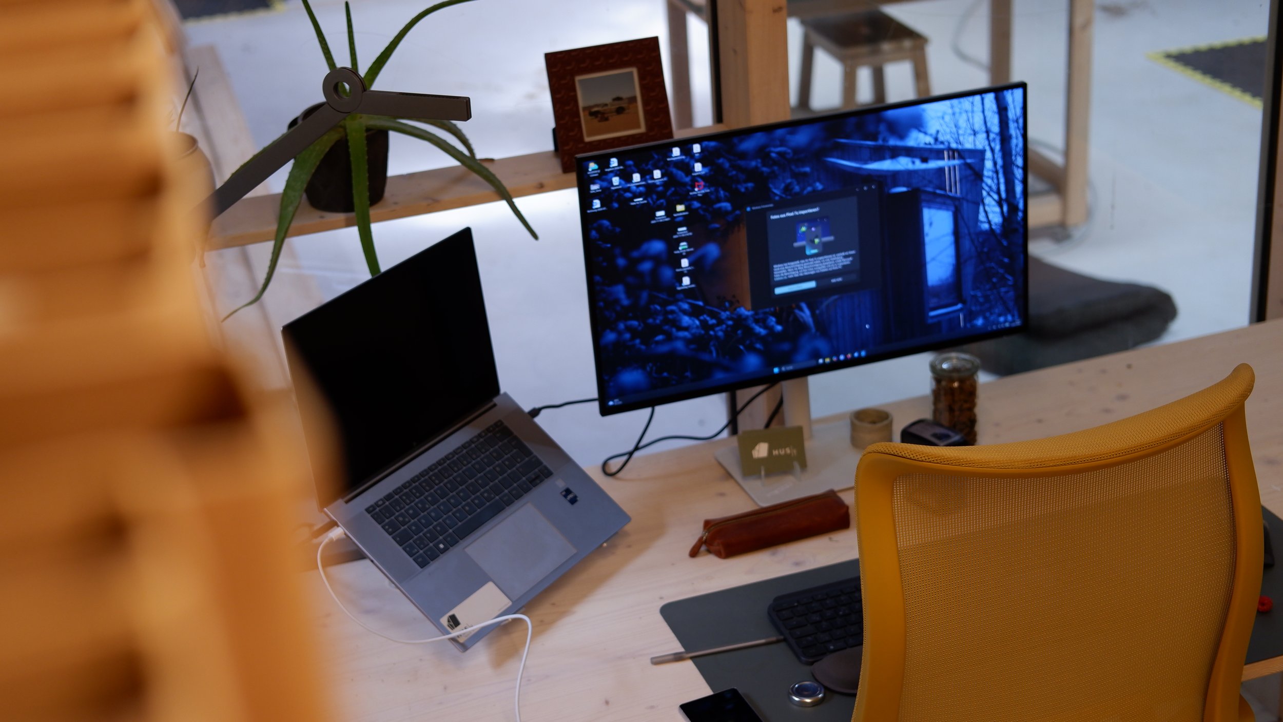 Ein Schreibtisch mit einem Laptop, einem zweiten Monitor, einer Tastatur, einer Maus und anderen Gegenständen. Im Hintergrund sind Holzregale und eine Pflanze. Es befindet sich eine gelbe Bürostuhl vor dem Tisch.