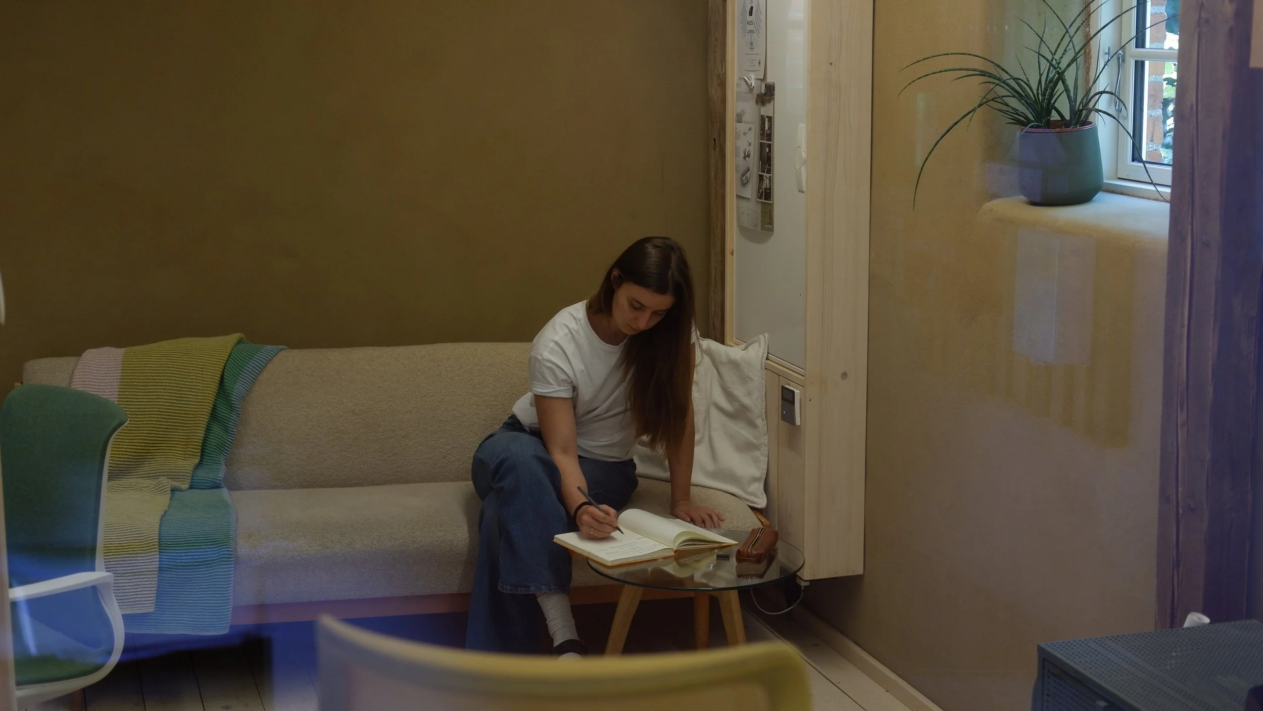 Eine junge Frau sitzt auf einem Sofa, liest in einem Buch und schreibt in ein Notizbuch. Hinter ihr an der Wand hängt eine weiße Wand mit Bildern oder Zeitschriften. Neben dem Sofa steht ein kleiner runder Tisch mit Brillenetui und einer Brille. Im Hintergrund ist ein Fenster mit einem grünen Blumentopf auf dem Fensterbrett. Das Zimmer ist gemütlich und modern eingerichtet.