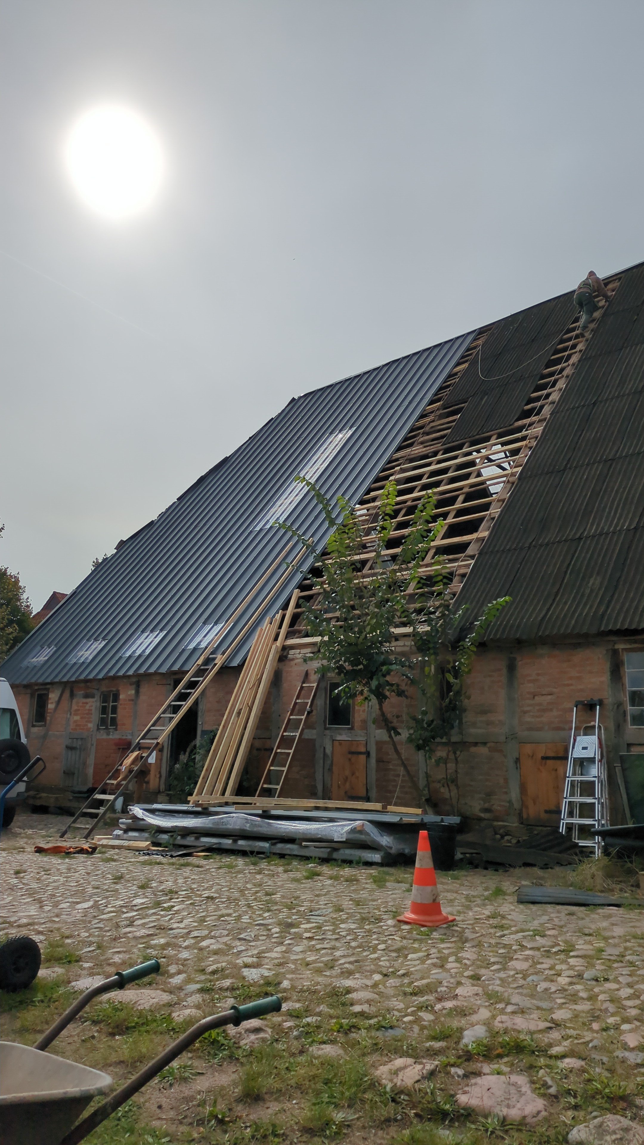 Ein Haus wird renoviert, mit Arbeitern auf dem Dach, das teilweise mit Metallplatten bedeckt ist. Der Himmel ist bewölkt, und im Vordergrund sind Baumaterialien, Werkzeuge und eine Markierung in Form eines orangefarbenen Verkehrscones zu sehen.