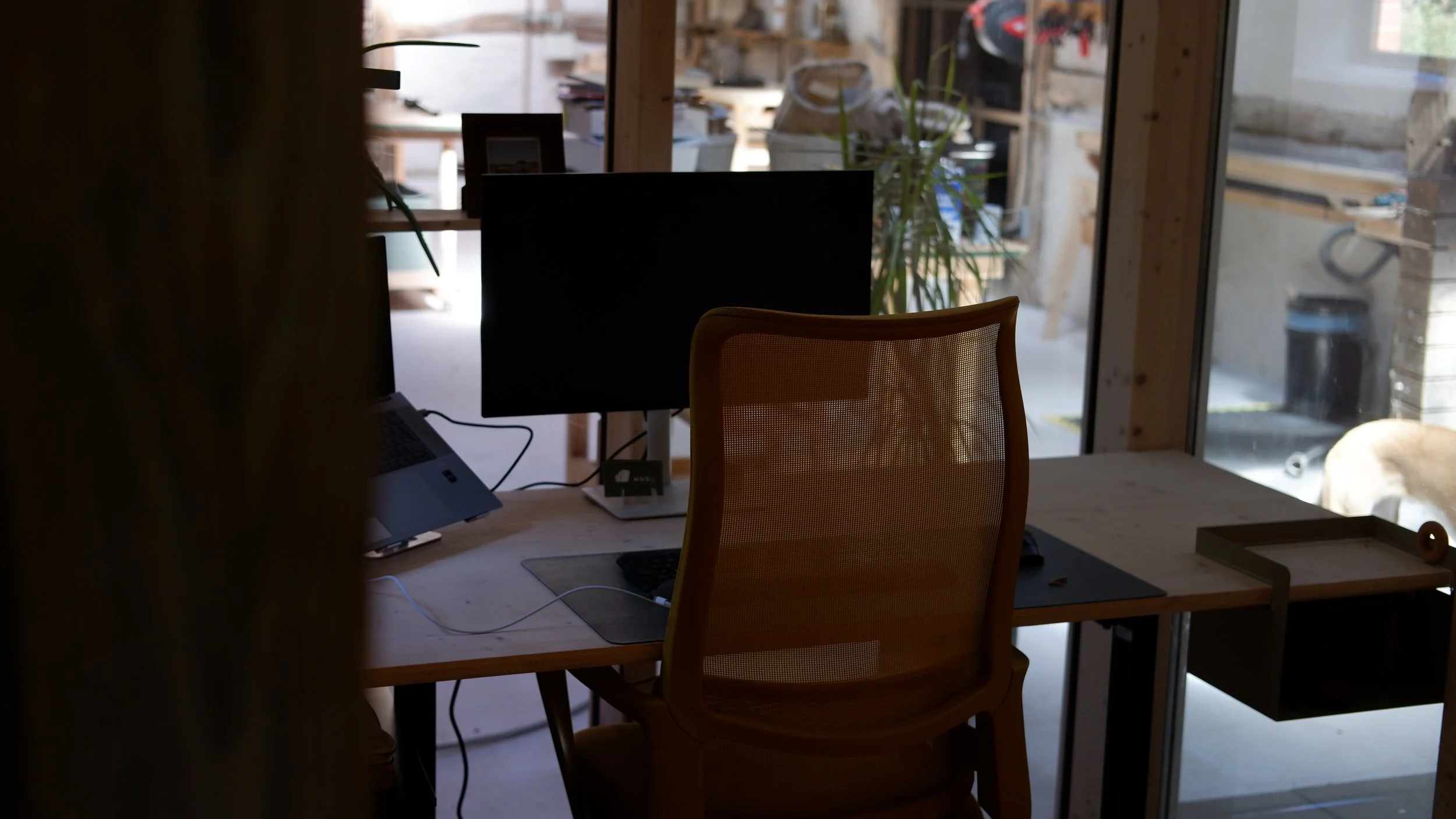 Ein dunkler Arbeitsbereich mit einem Tisch, einem Computerbildschirm, einer Tastatur und einem Telefon, im Hintergrund Fenster mit Gartenansicht, Pflanzen und Werkzeuge im Außenbereich.