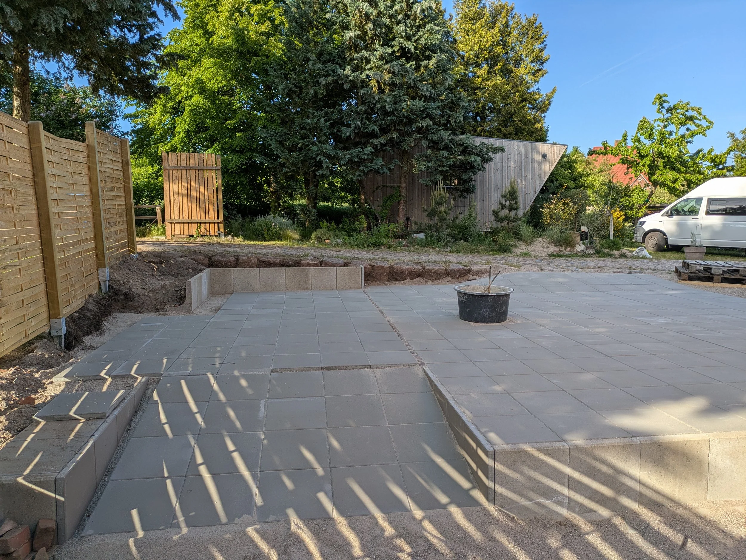Der Garten im Bau mit Pflastersteinen, einem Eimer und Zäunen, umgeben von Bäumen und einem blauen Himmel.