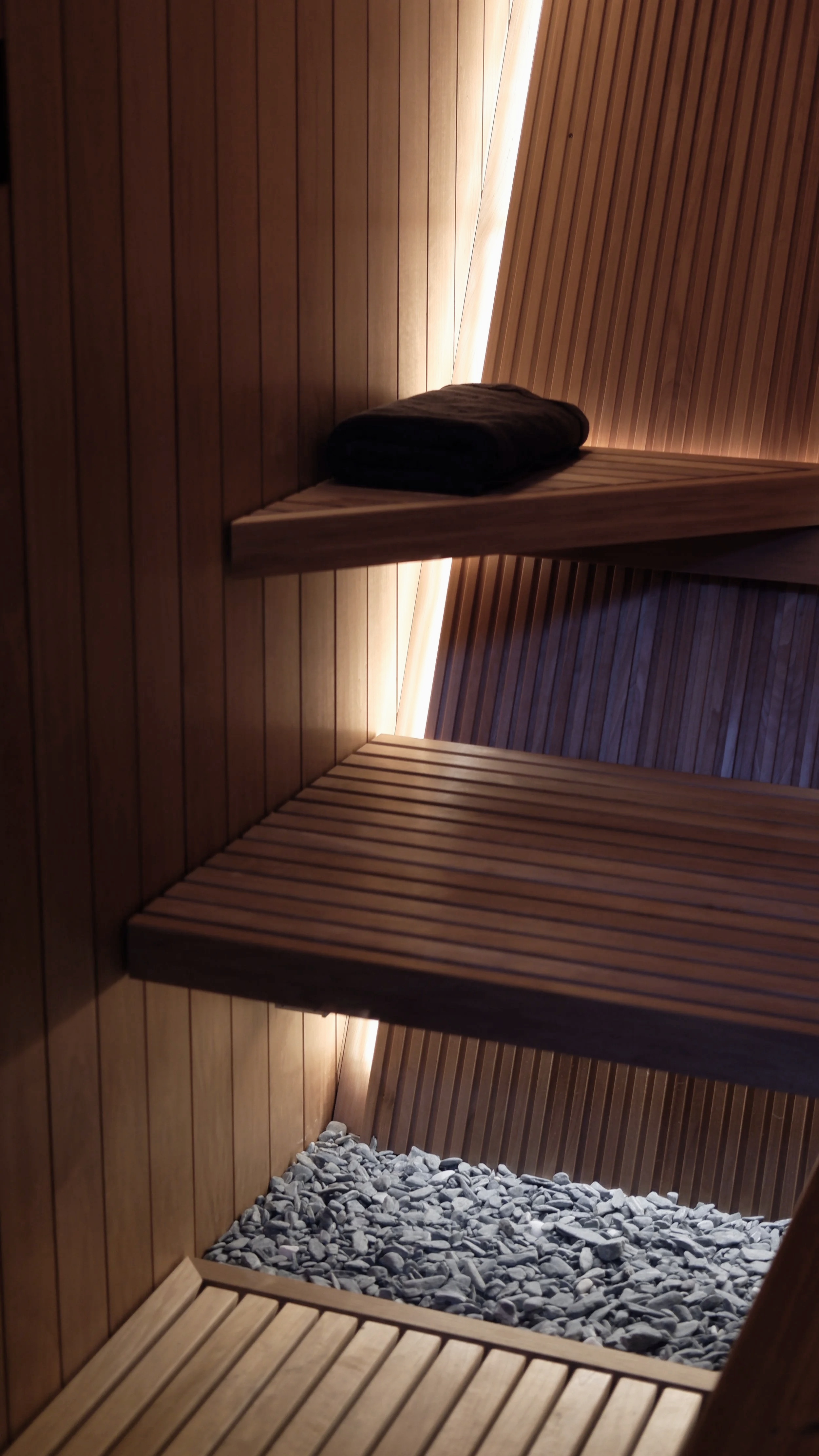Innenraum einer Sauna mit Holzverkleidung, einem gefalteten Handtuch auf einer Bank, Steinen auf dem Boden und indirekter Beleuchtung.
