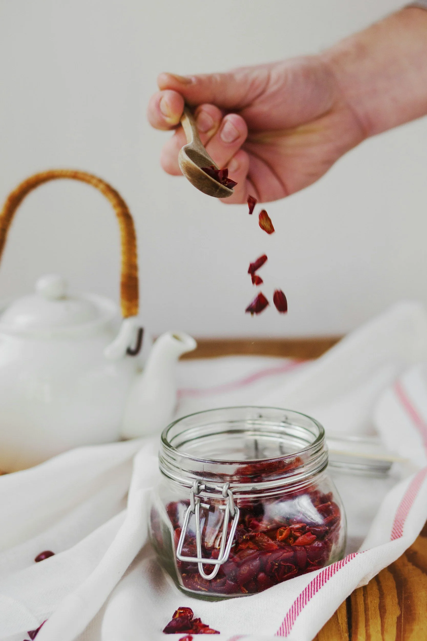 Hand sprinkling dried cranberries into a glass jar on a table with a white teapot and towel.