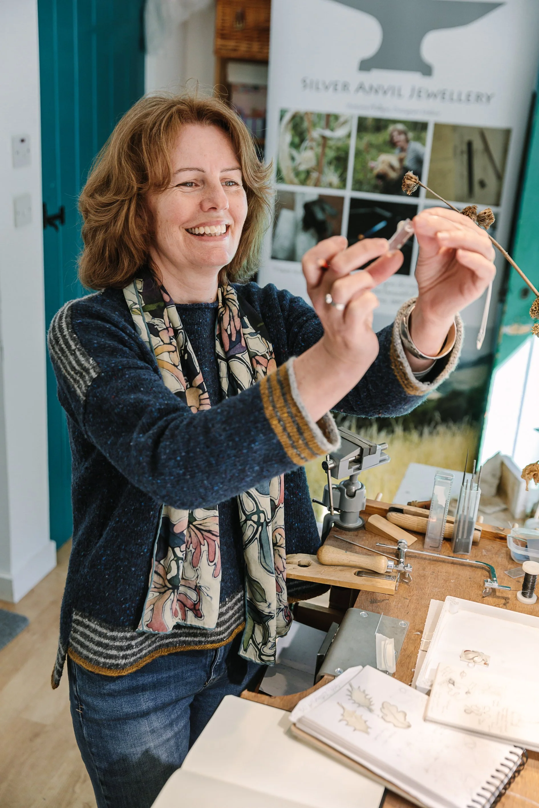 Person in a sweater and scarf crafting jewelry at a workbench, surrounded by tools and sketches, with a "Silver Anvil Jewellery" banner in the background.