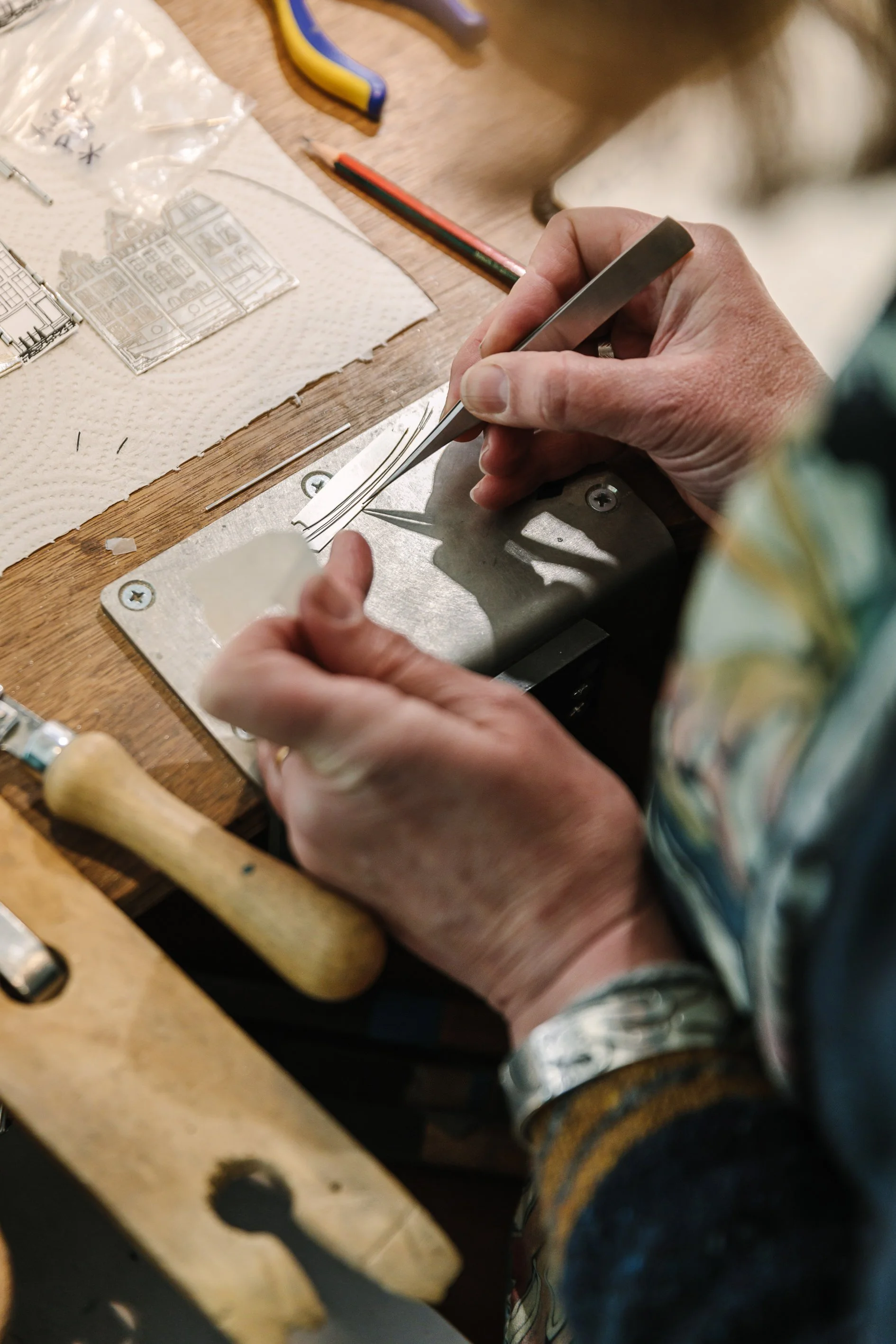 Victoria working with metal on a wooden table, holding tweezers and small metal pieces, with tools and drawings nearby.