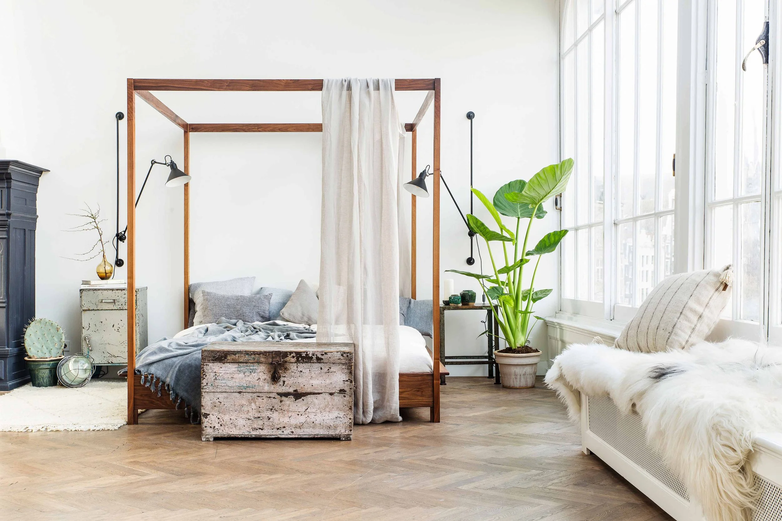 A bright bedroom with a four-poster bed, white walls, large windows, and indoor plants.