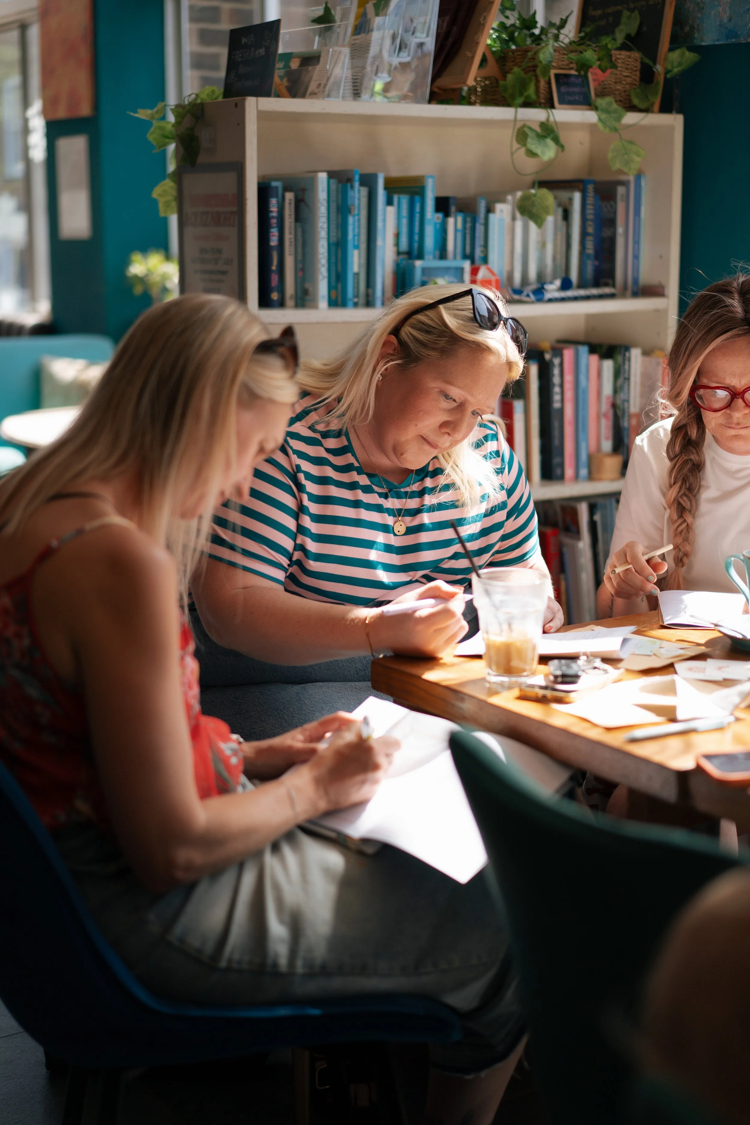 Three women sitting at a wooden table, reading and writing, with bookshelves filled with books and plants in the background.