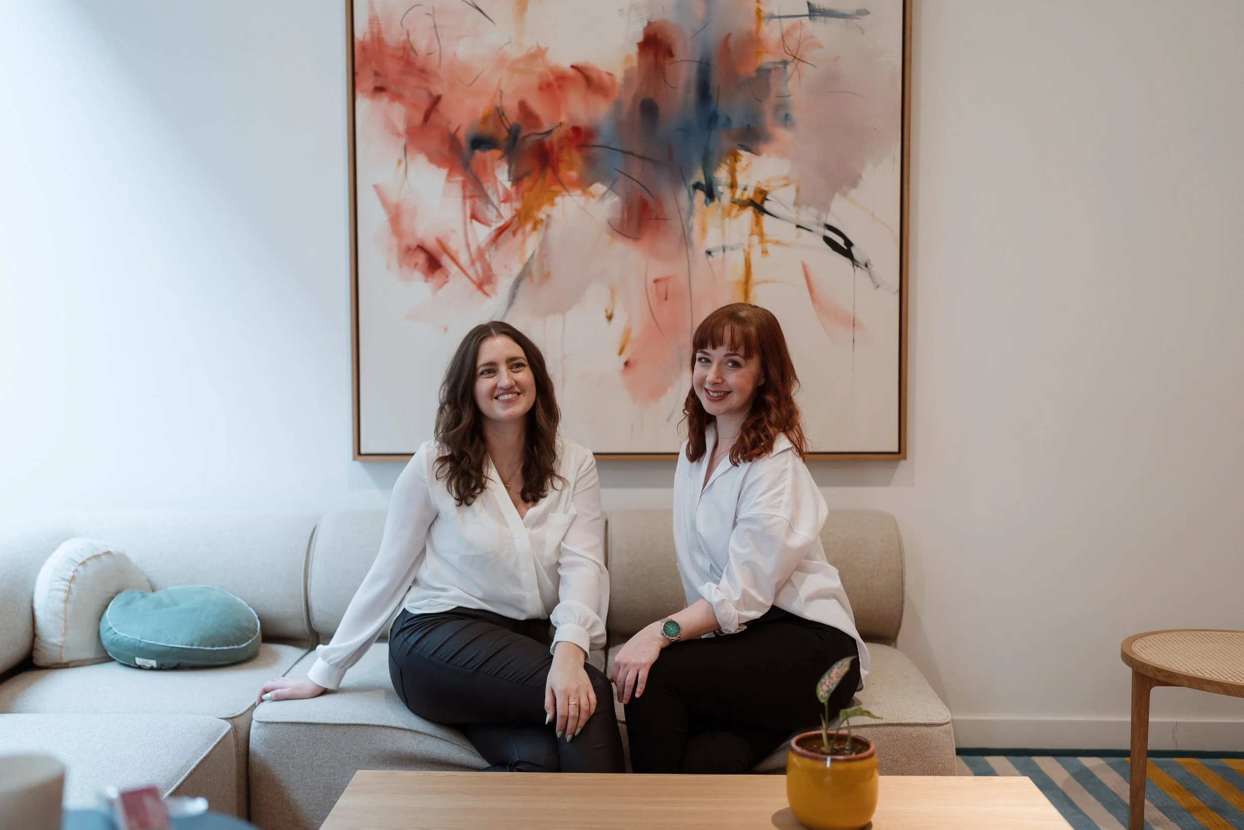 Two women sitting on a beige sofa in a modern living room, smiling and wearing white blouses. Behind them is an abstract painting with splashes of red, blue, and black. A small wooden table with a yellow pot containing a small plant is in the foreground.