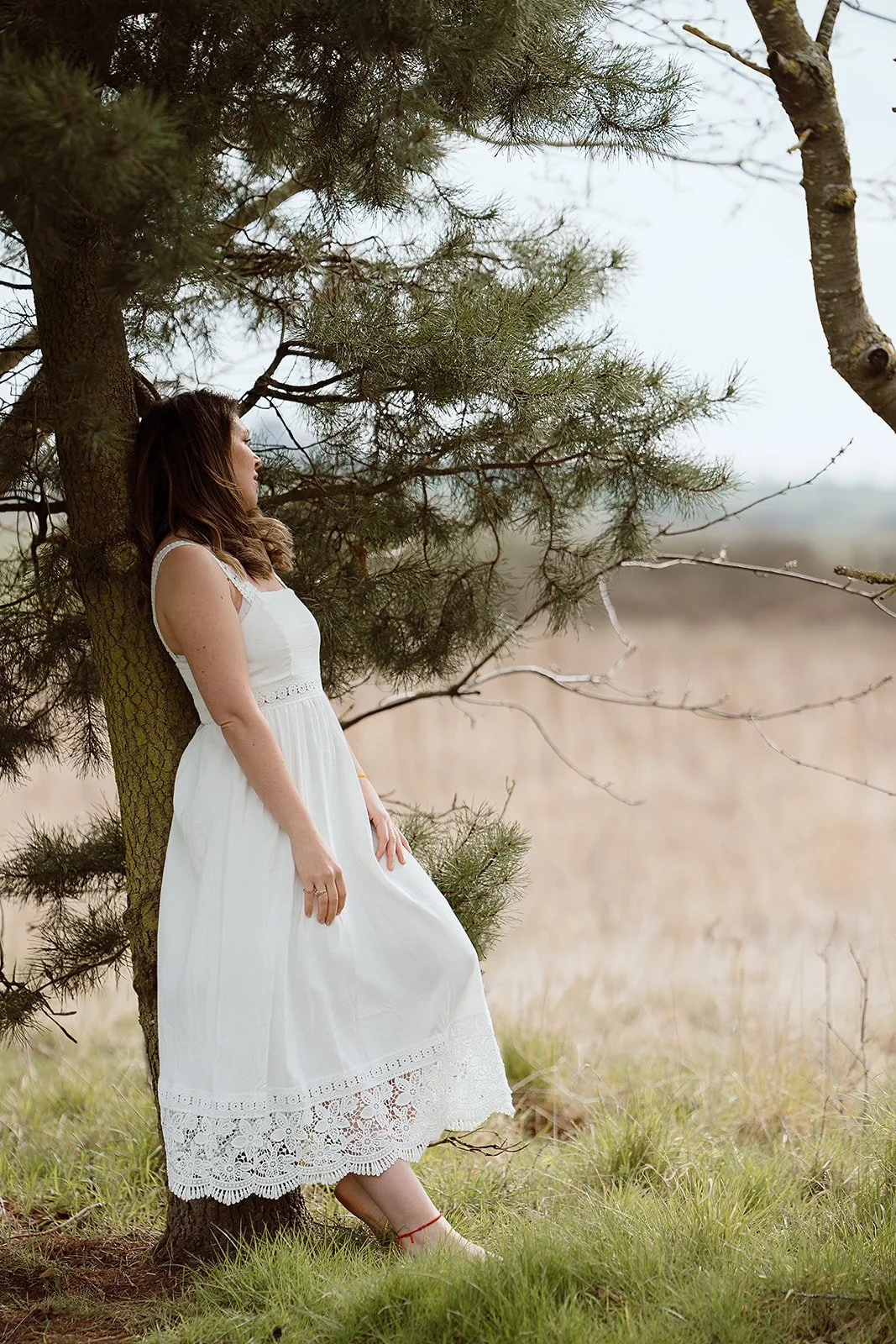 A woman in a white dress leaning against a tree in a field with tall grass and overcast sky.