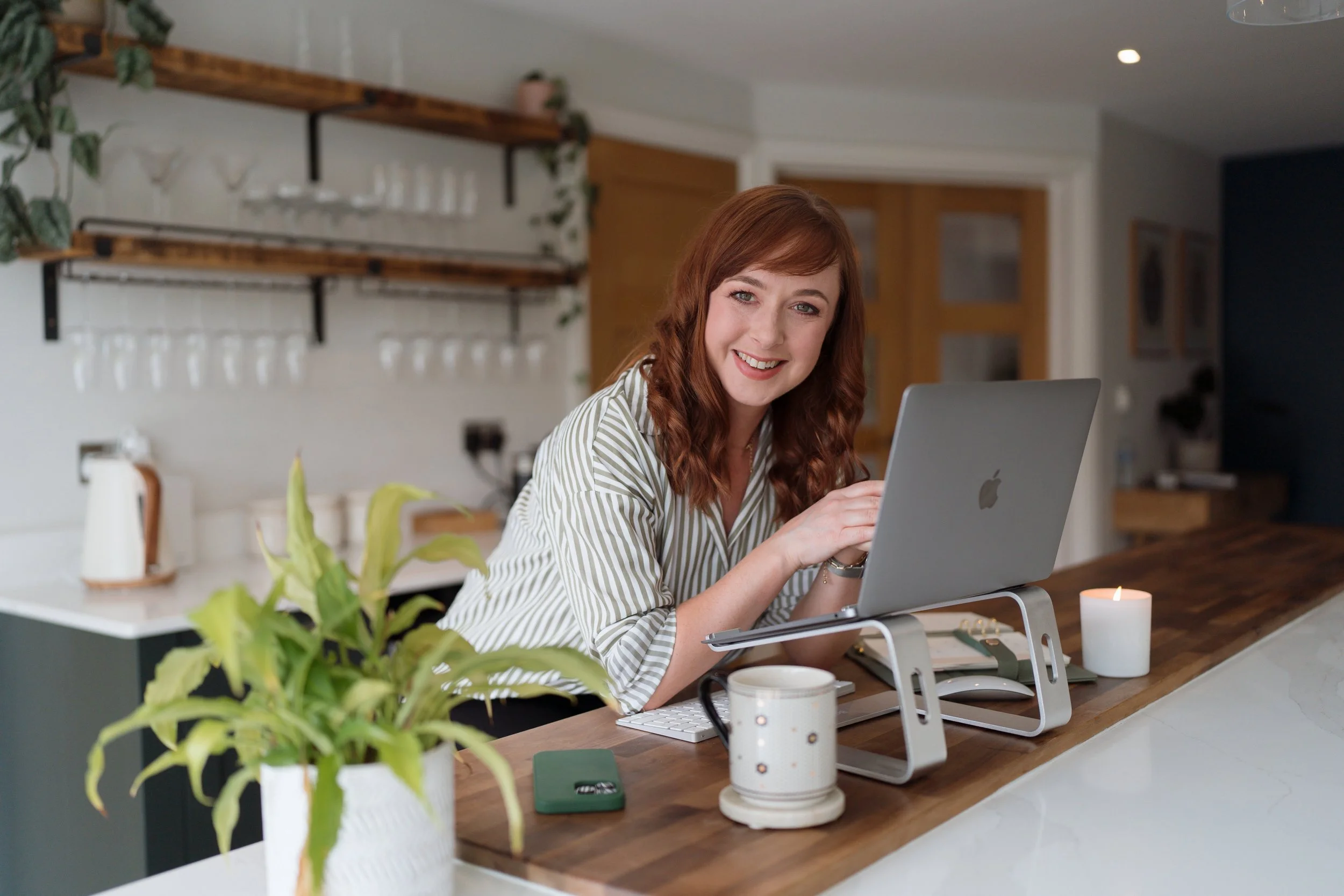 A woman with red hair smiles while working on a laptop at a kitchen counter with a plant, mug, candle, and phone holder.