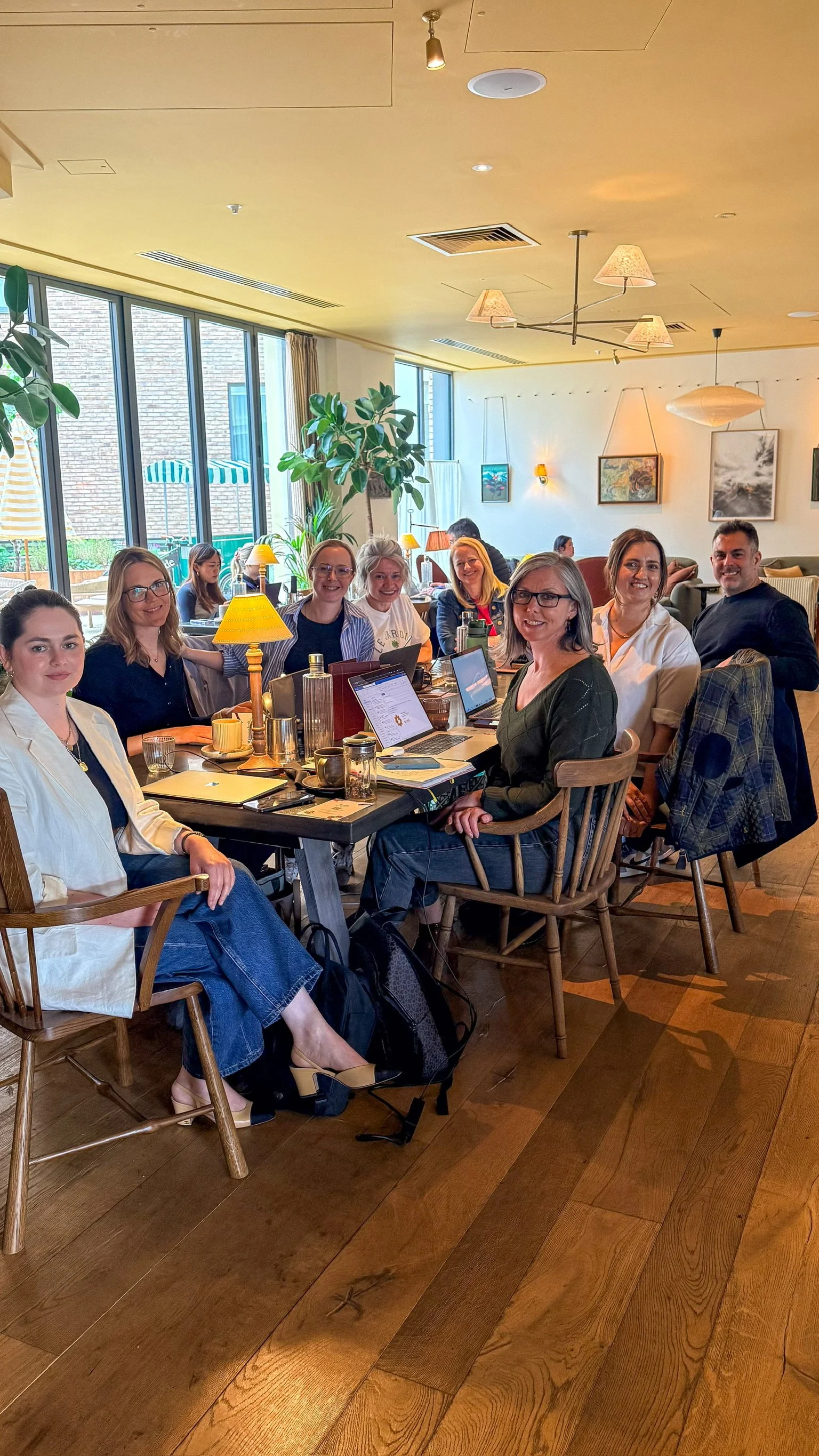 A group of approximately eight women and one man sitting around a table in a cozy, well-lit restaurant or cafe. The table has laptops, notebooks, and drinks. Large windows, green plants, and framed artwork decorate the background. The ceiling has warm lighting fixtures and hanging lamps.