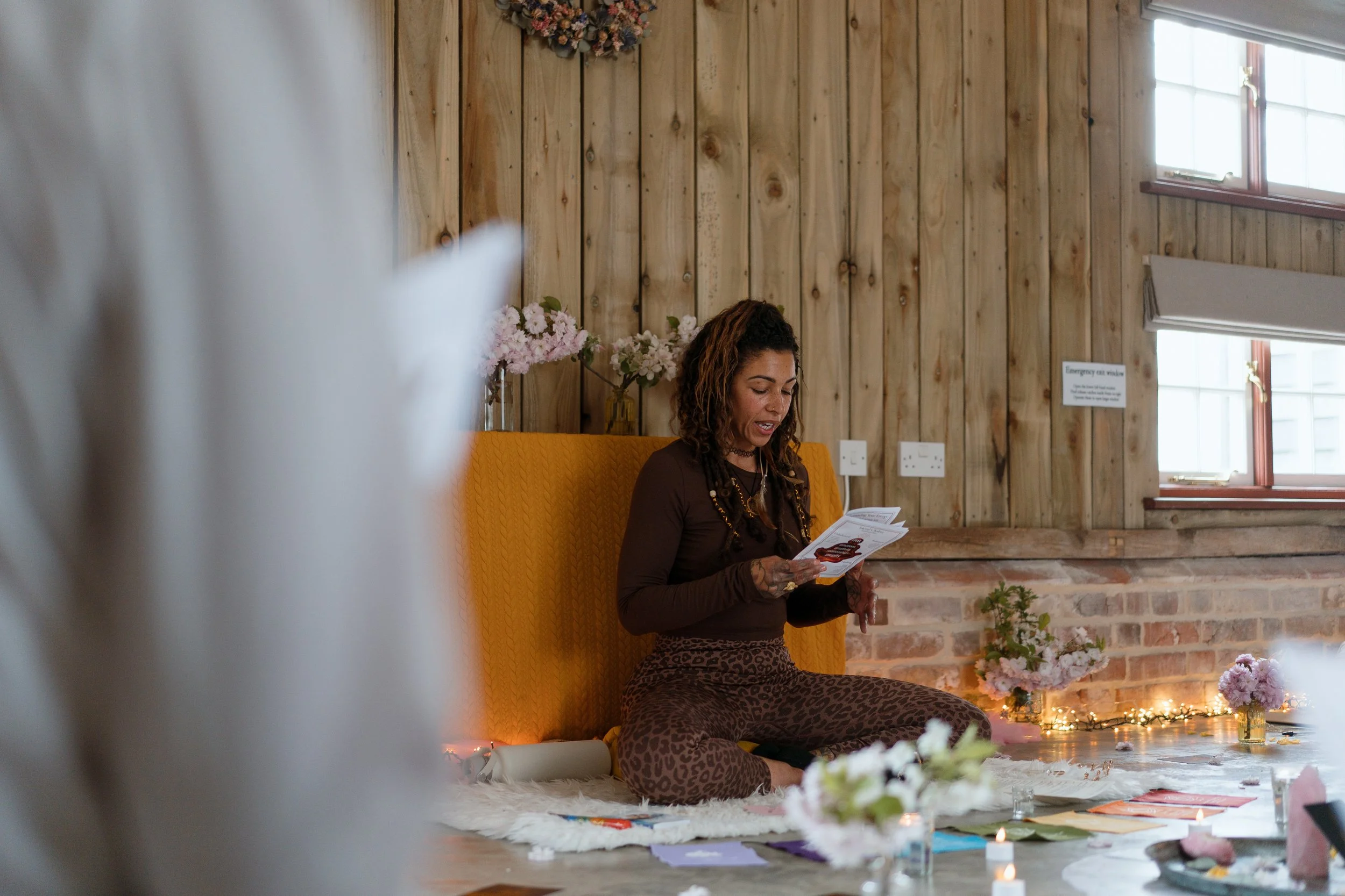 A woman with tattoos and long curly hair sitting cross-legged on a white rug, reading a booklet, in a cozy room with wood-paneled walls and flower decorations.