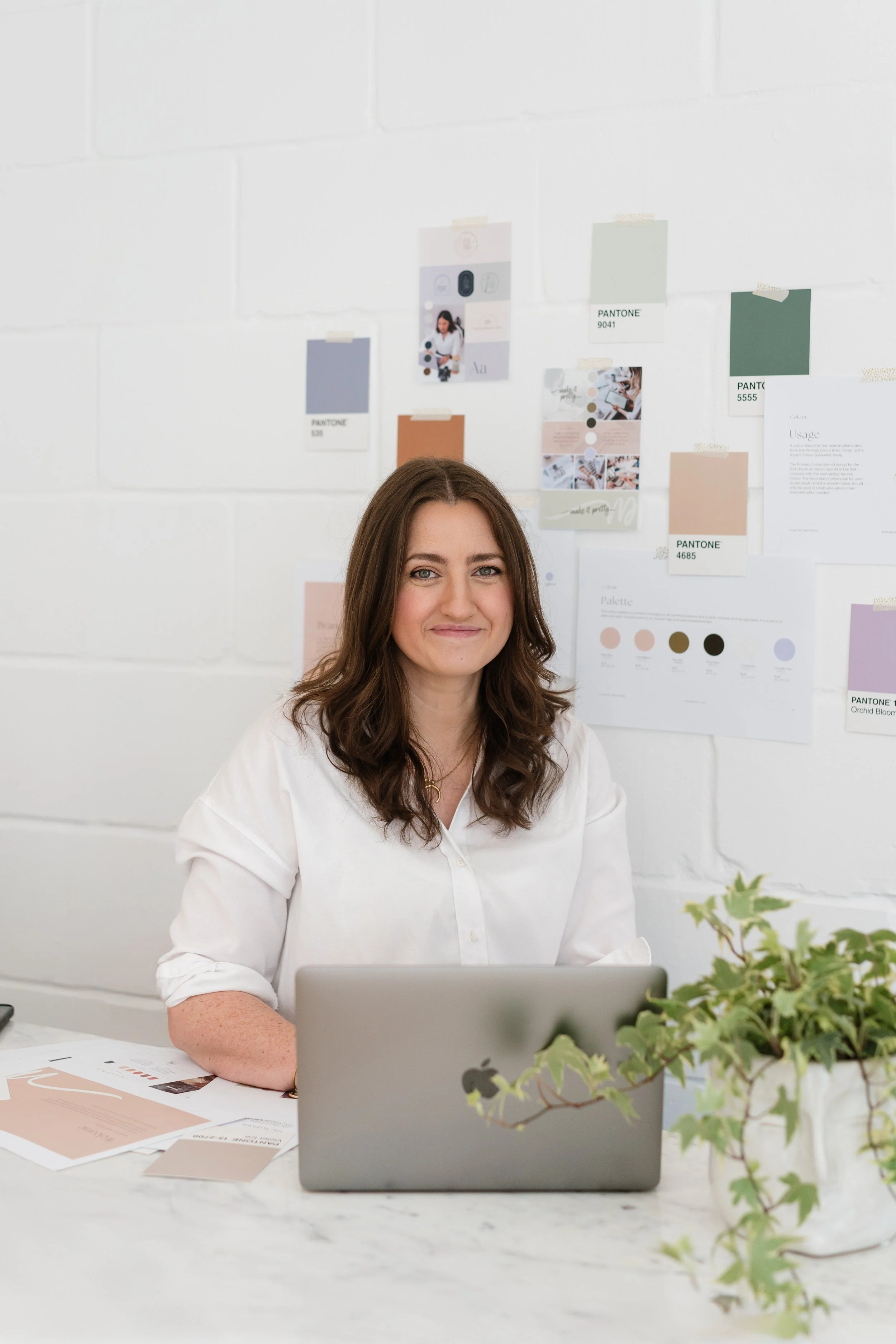 A woman with long brown hair sitting at a white desk with a silver laptop in front of her, papers and brochures on the desk, and a potted green plant on the right side, in a white-walled office with color swatches and design samples on the wall.
