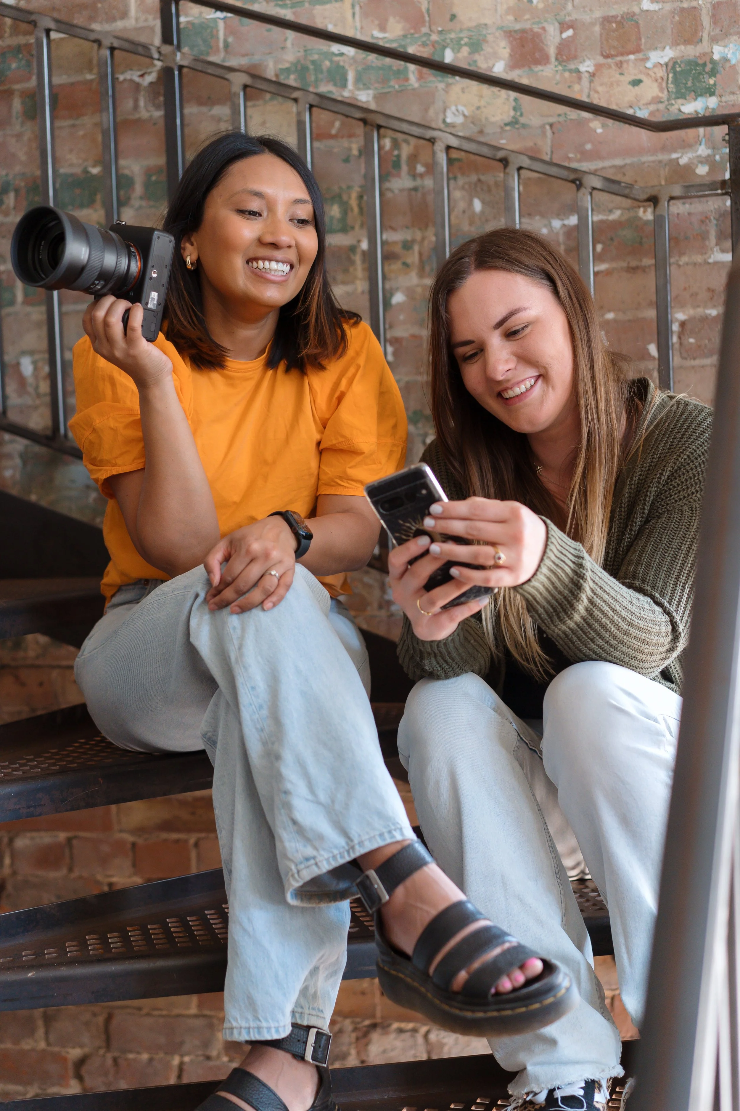 Two young women sitting on a staircase with an exposed brick wall behind them, one holding a camera and looking at her phone, smiling.