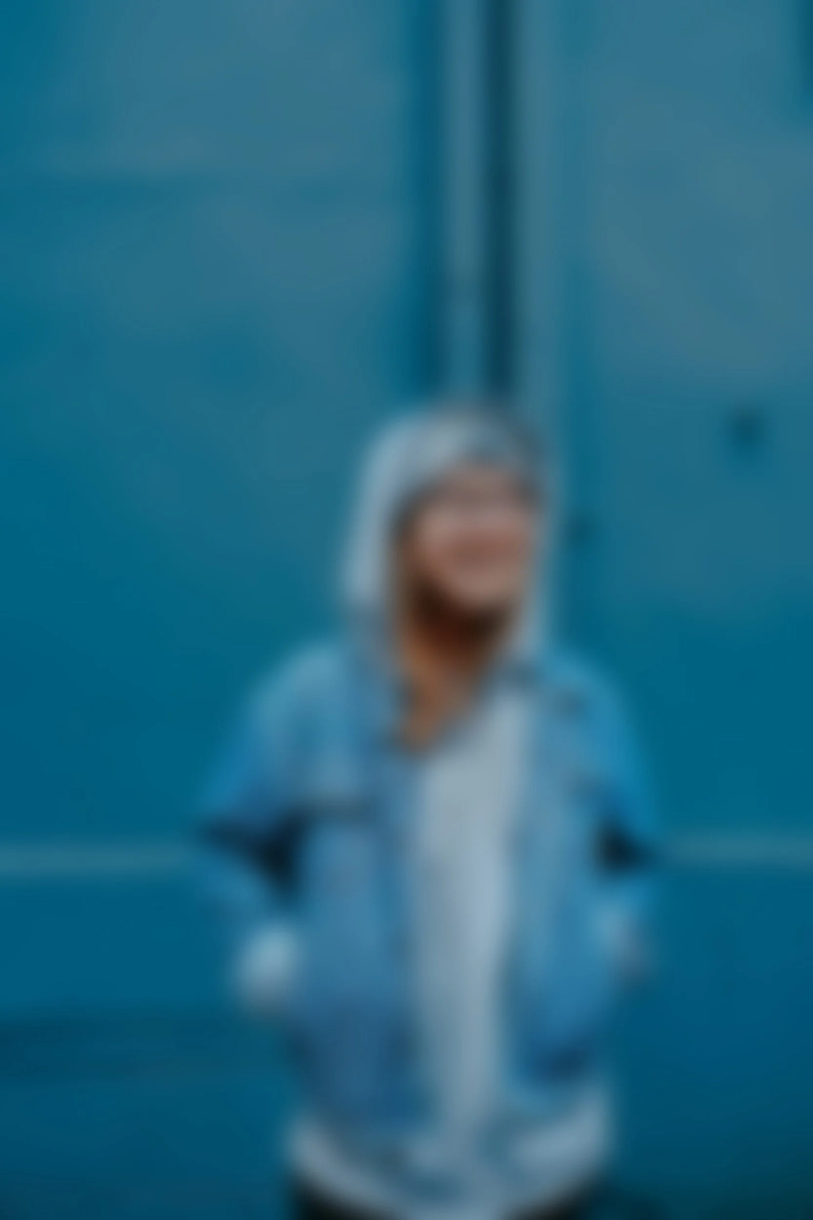 A person with short, gray hair wearing a gray hoodie and glasses, standing outdoors against a blue wall, smiling.